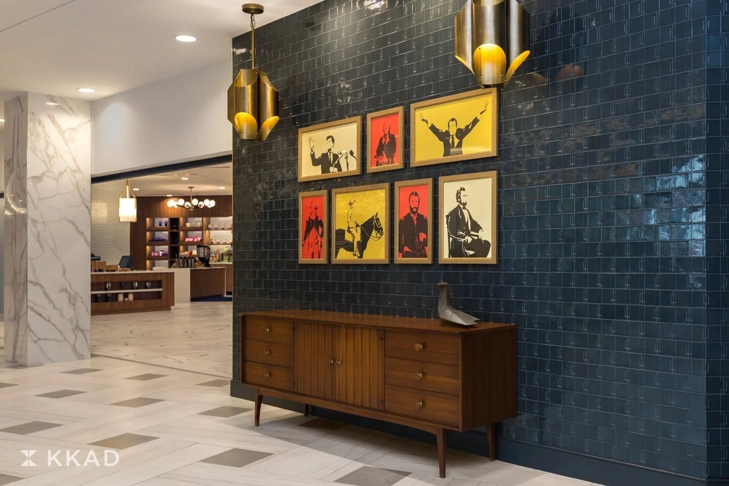 Interior of a modern building lobby featuring black tiled wall with framed pop art style portraits of musicians, wooden sideboard with a decorative bird sculpture, and gold pendant lights.