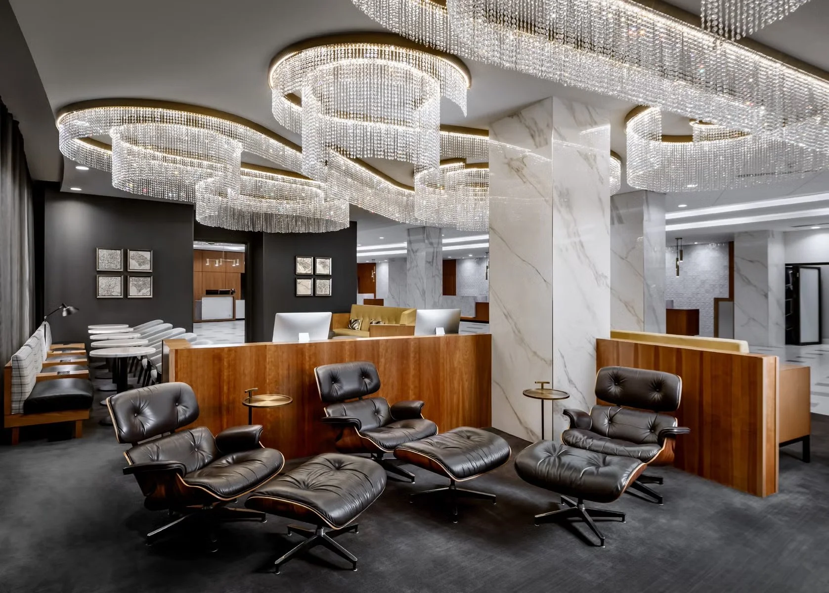 A luxurious hotel lobby featuring elegant chandeliers, marble columns, a sitting area with black leather lounge chairs, and a reception desk in the background.