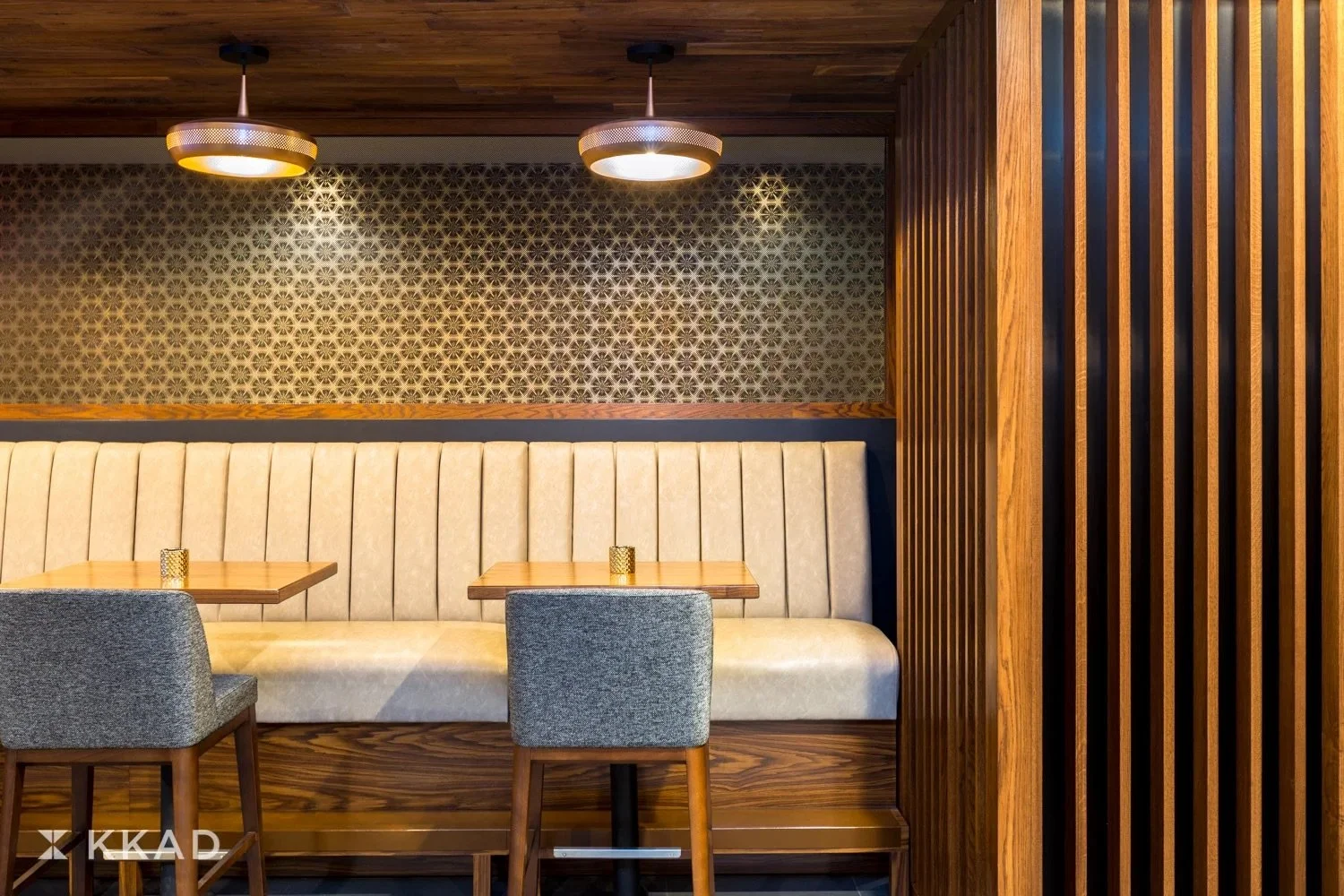 Interior of a restaurant with beige upholstered banquette seating, wooden tables, grey chairs, patterned wallpaper, and wooden wall paneling. Two ceiling lights are visible above.