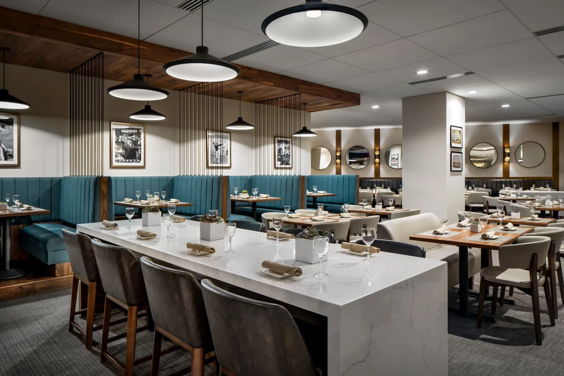 Modern restaurant with neatly set tables, white marble top, wine glasses, cloth napkins, and small potted plants, featuring a mix of blue, beige, and wooden accents, black and white framed photos on the walls, circular mirrors, and stylish pendant li