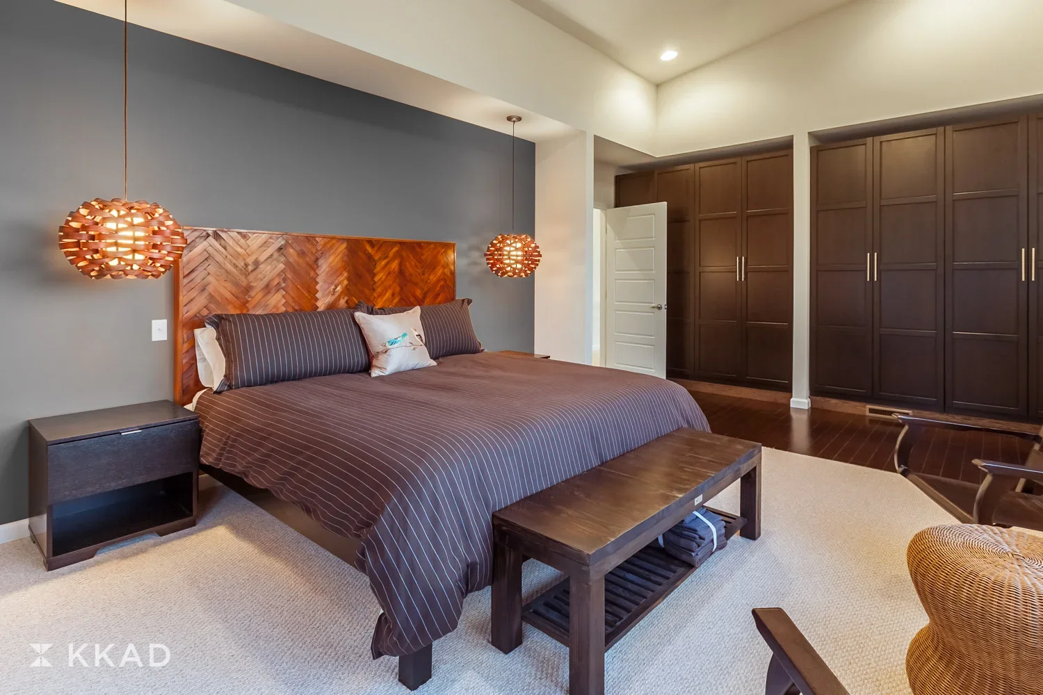 Modern bedroom with a large bed featuring a wooden headboard, dark striped bedding, and decorative pillows. Hanging pendant lights on both sides of the bed, dark wooden nightstand, and built-in dark wood closet. Light-colored walls and a white door. 