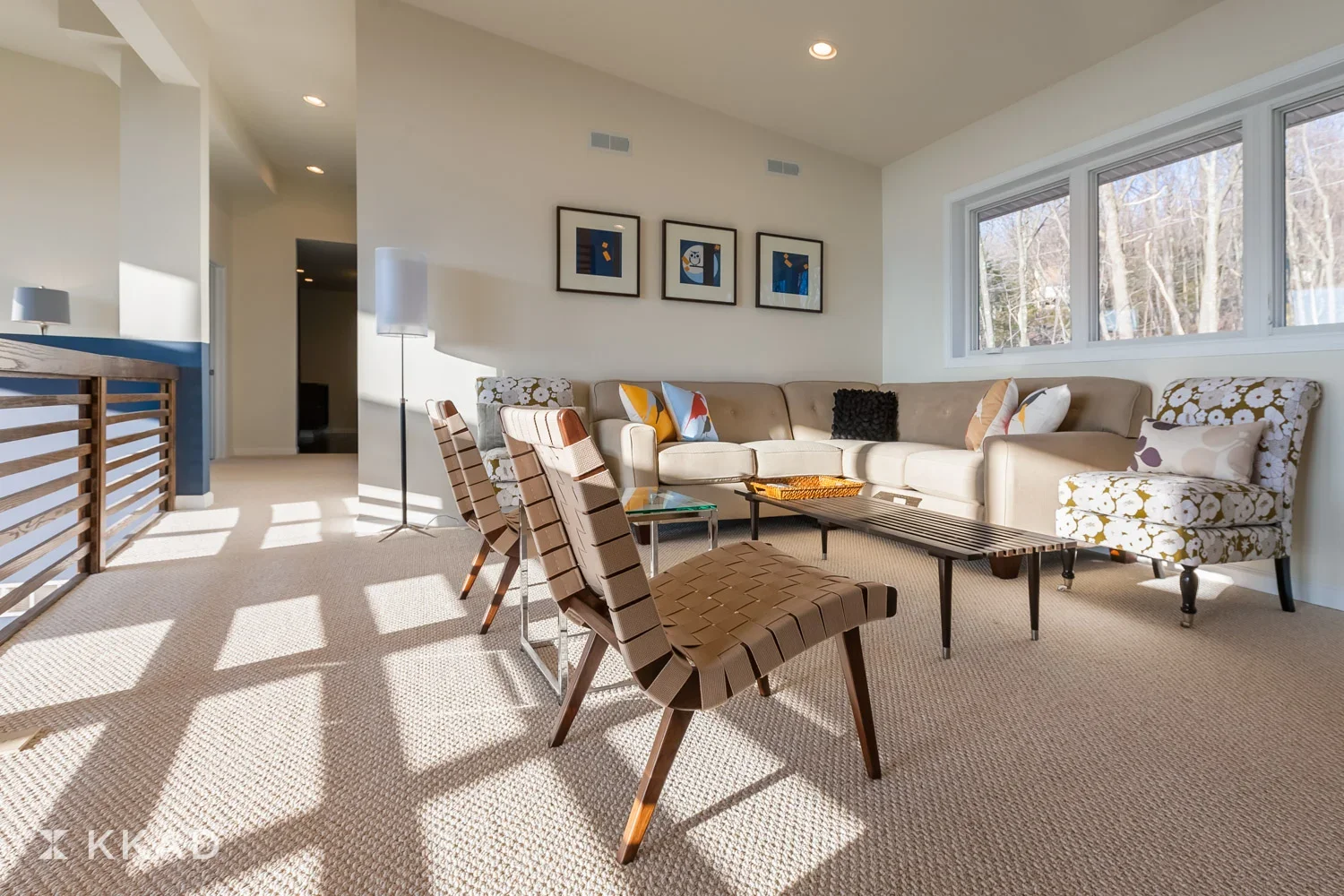 Bright living room with beige sofa, patterned armchairs, four framed pictures, large window showing trees, and wood coffee tables.