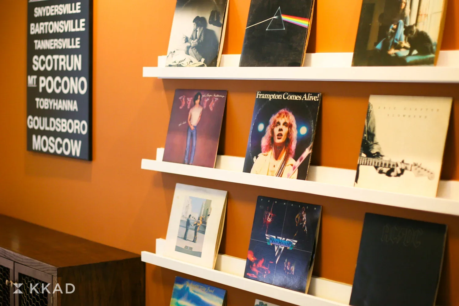 Display of vinyl records on white shelves against an orange wall, featuring album covers including Pink Floyd and other artists.