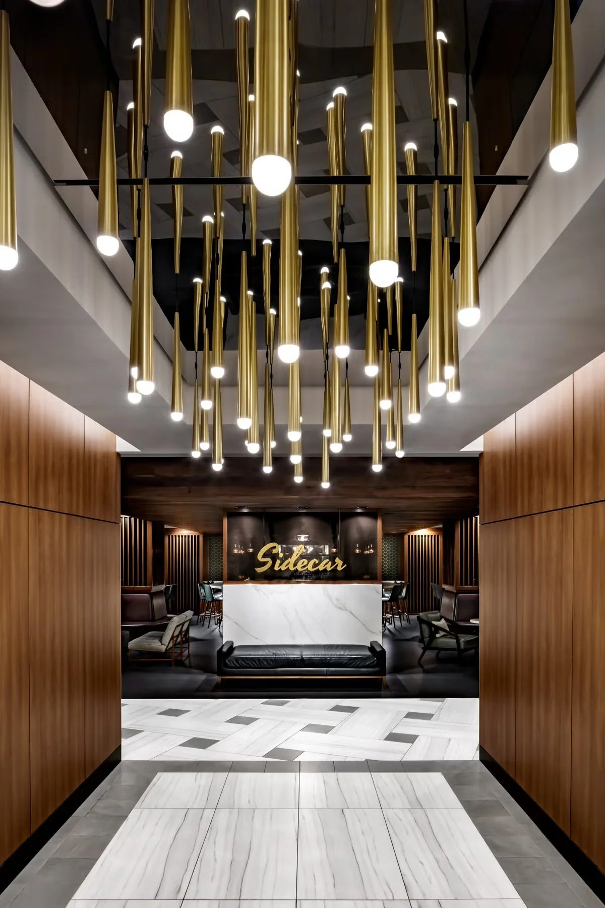 Modern hotel lobby featuring a chandelier with multiple vertical gold rods, wooden wall panels, a white and gray marble floor, and seating areas on either side. A reception desk with a sign that reads 'Sidecar' is visible in the background.
