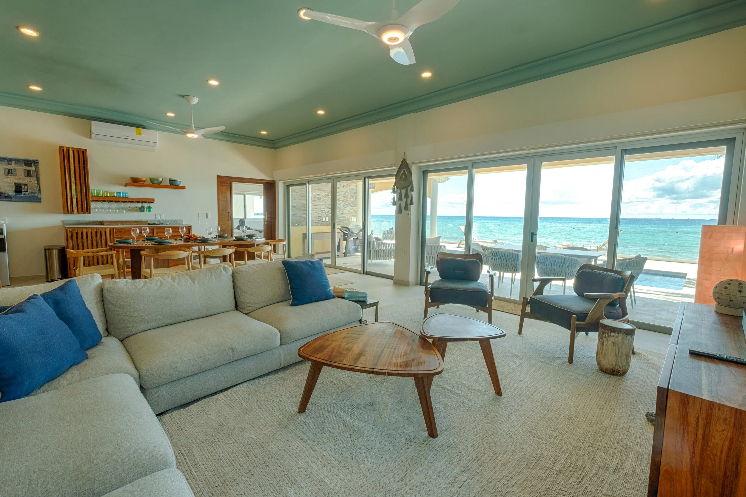 Living room with a view of the ocean, featuring a light-colored sofa with blue pillows, wooden chairs, a coffee table, and large sliding glass doors leading to a balcony with seating.