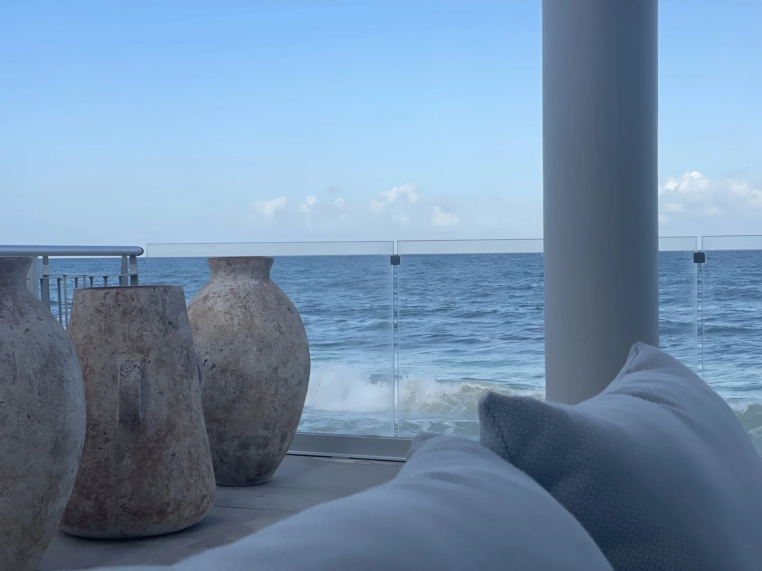 View of the ocean with blue sky and a few clouds, taken from a terrace with large ceramic pots and a glass railing, and part of a white sofa with a pillow in the foreground.