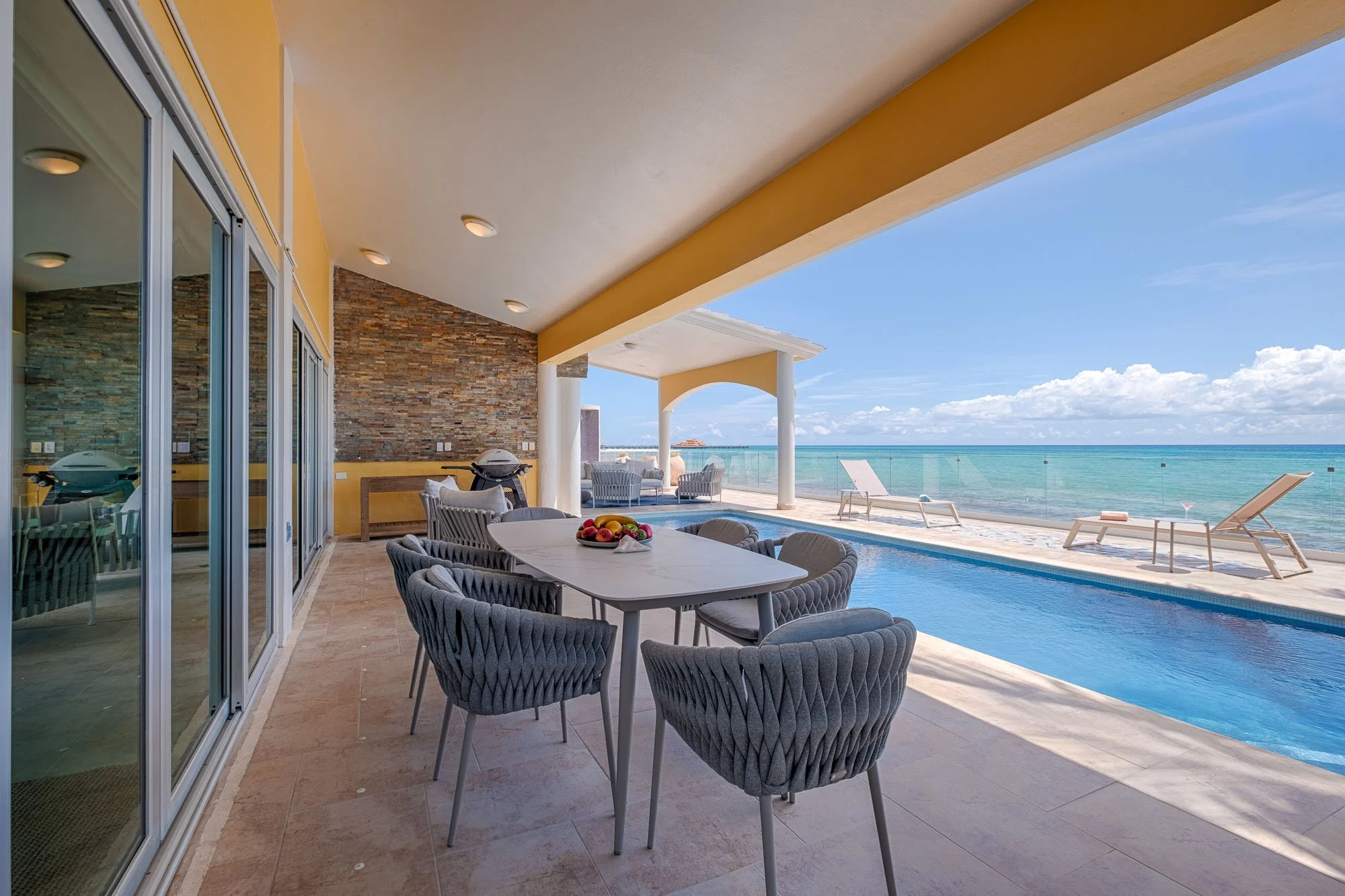 Outdoor patio with dining table and chairs, view of a swimming pool and the ocean with lounge chairs, under a covered patio area.