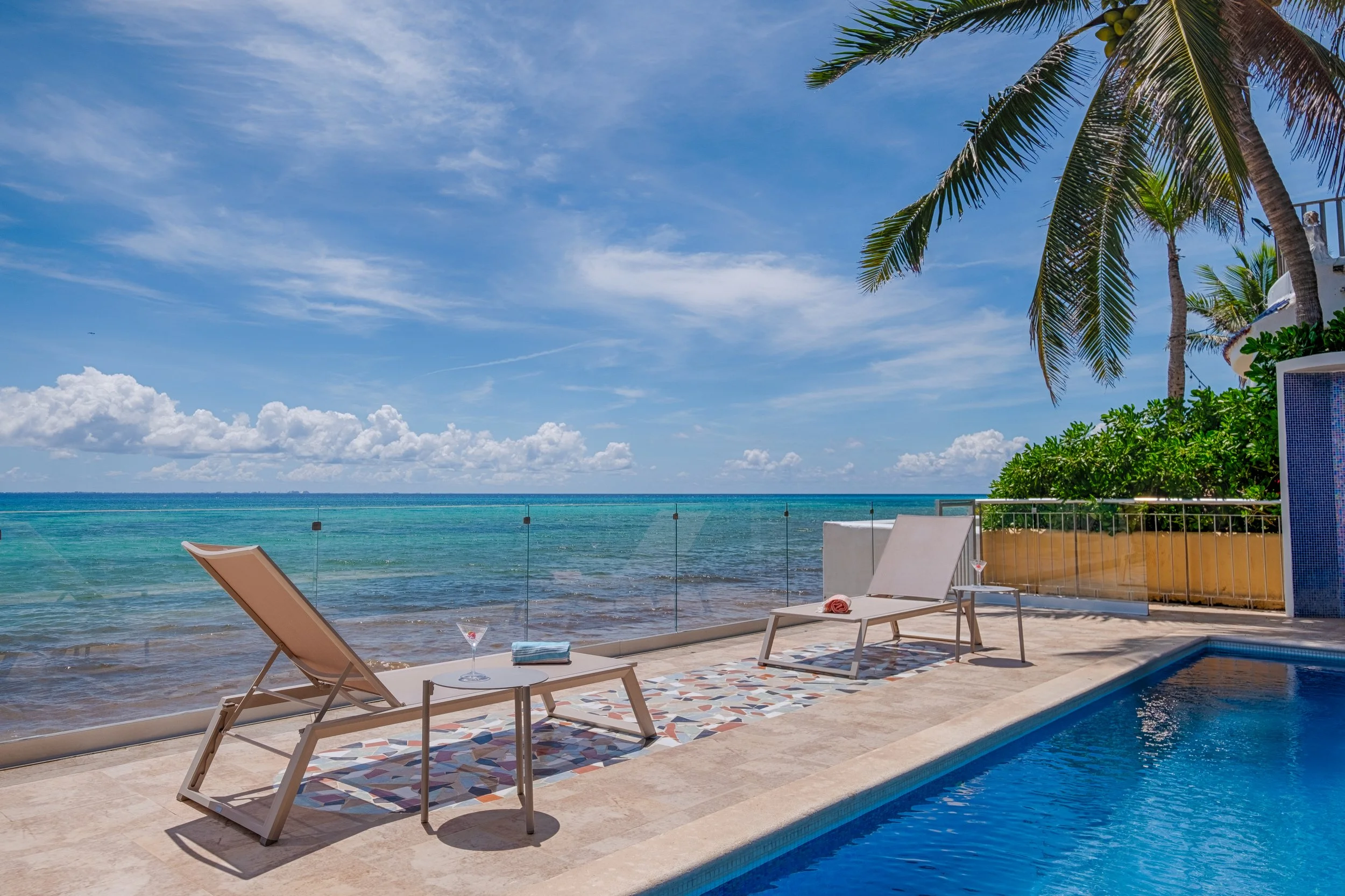 Ocean view with lounge chairs and a pool on a sunny day with blue sky and palm trees.