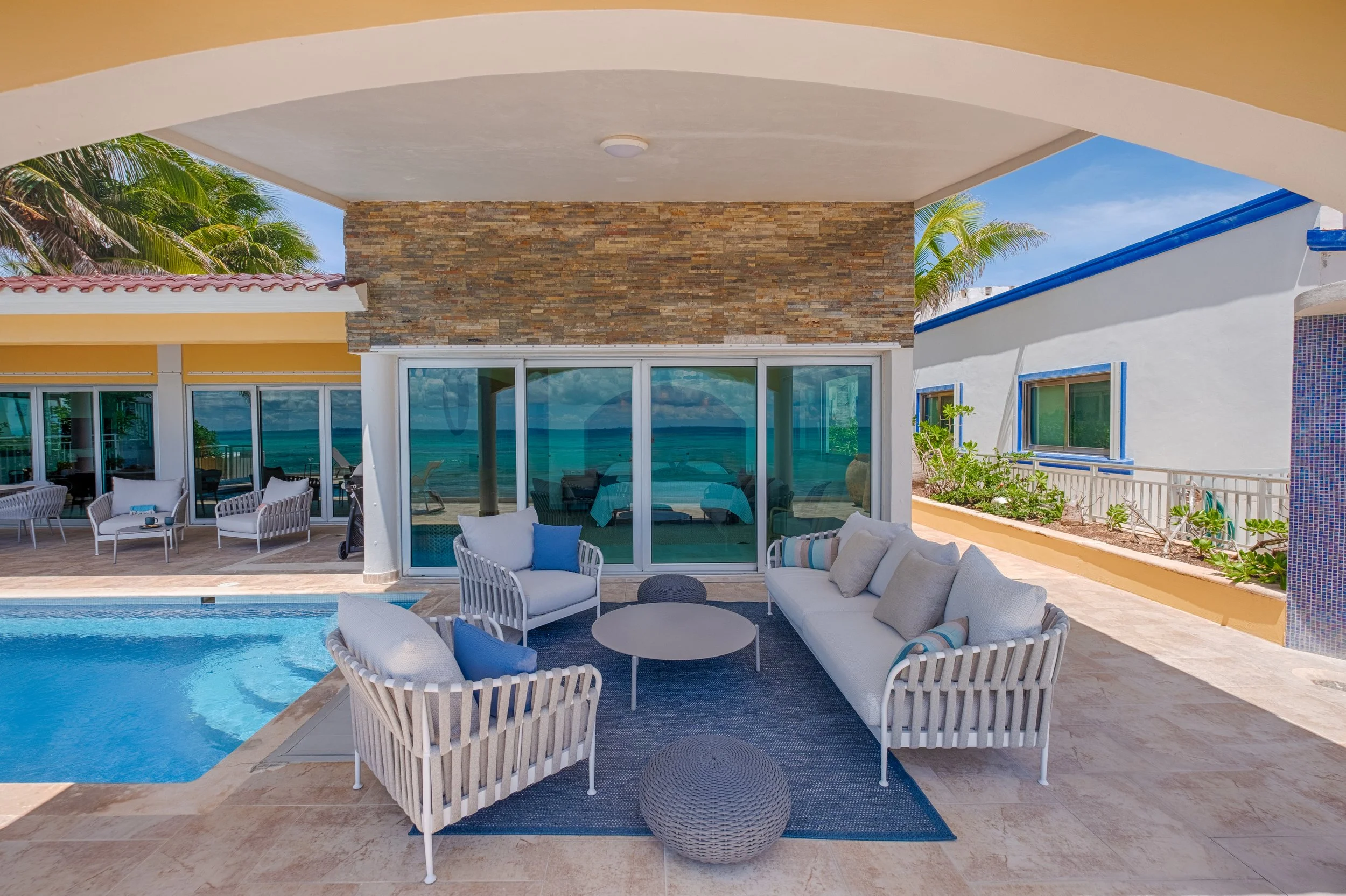 Outdoor patio with seating area, including a white sofa, armchairs, and a round coffee table, next to a swimming pool. Behind, sliding glass doors lead to an indoor space with ocean view. Surrounding the patio, there are tropical plants and a white b