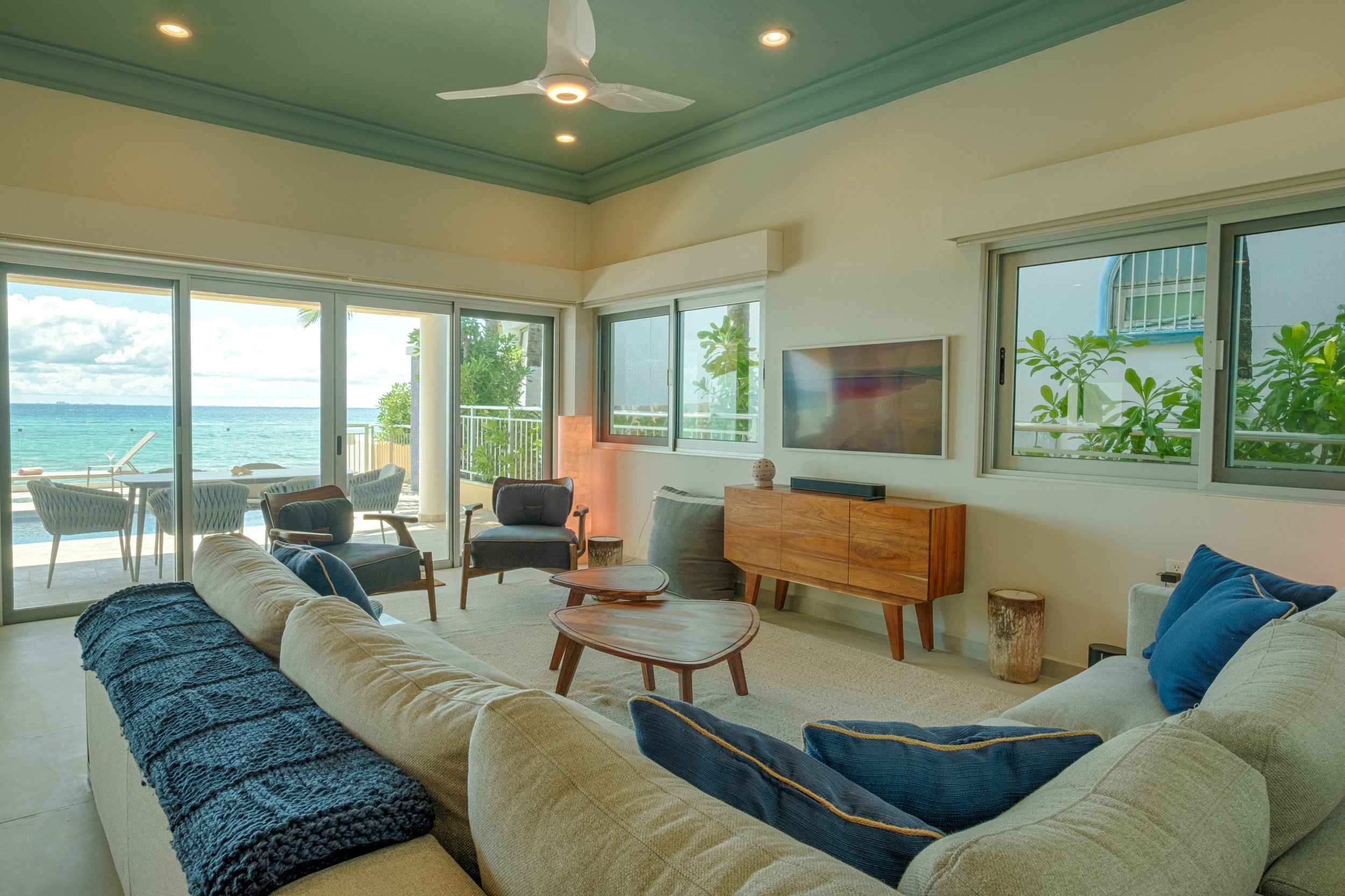 Living room with large glass sliding doors overlooking the beach, featuring beige sofas with blue pillows, wooden coffee tables, and a wooden TV stand, with a ceiling fan and tropical plants visible outside.