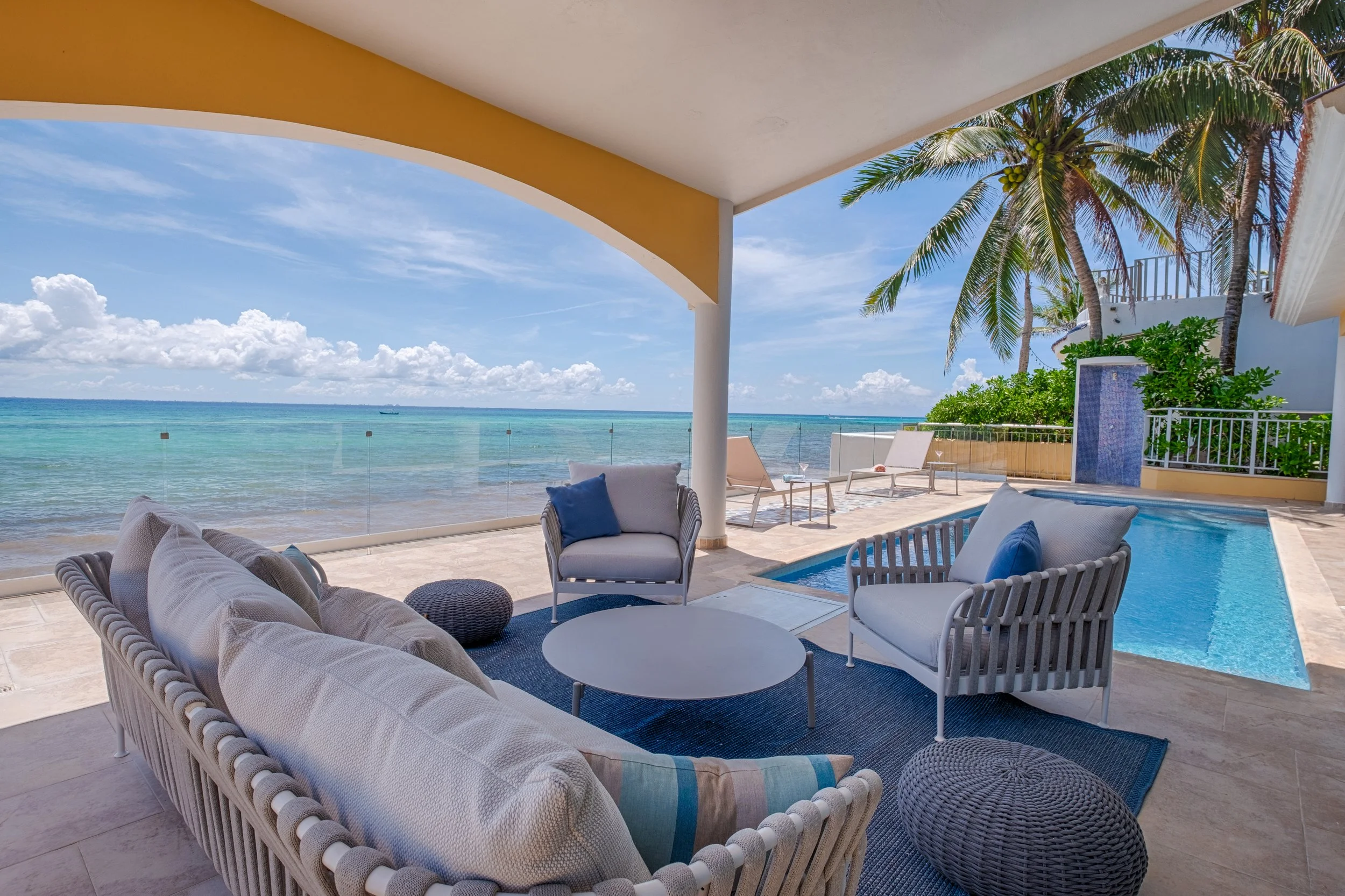 A covered patio with outdoor seating, a small pool, and an ocean view featuring blue sky, white clouds, and palm trees.