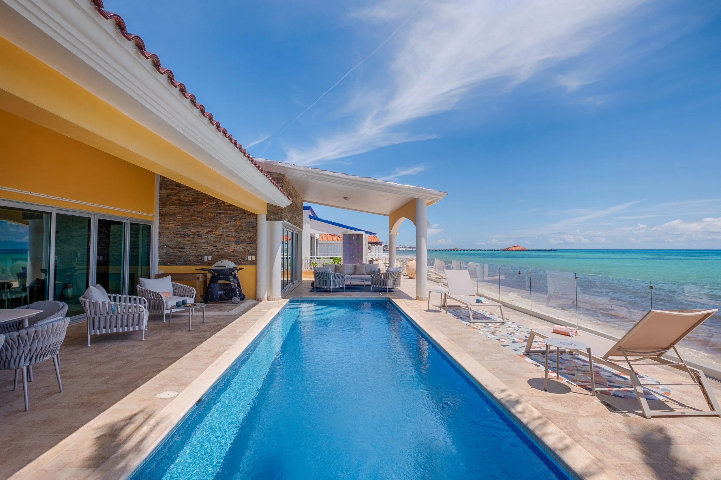 Poolside view at a beach resort with lounge chairs, a glass railing, ocean, and clear blue sky.