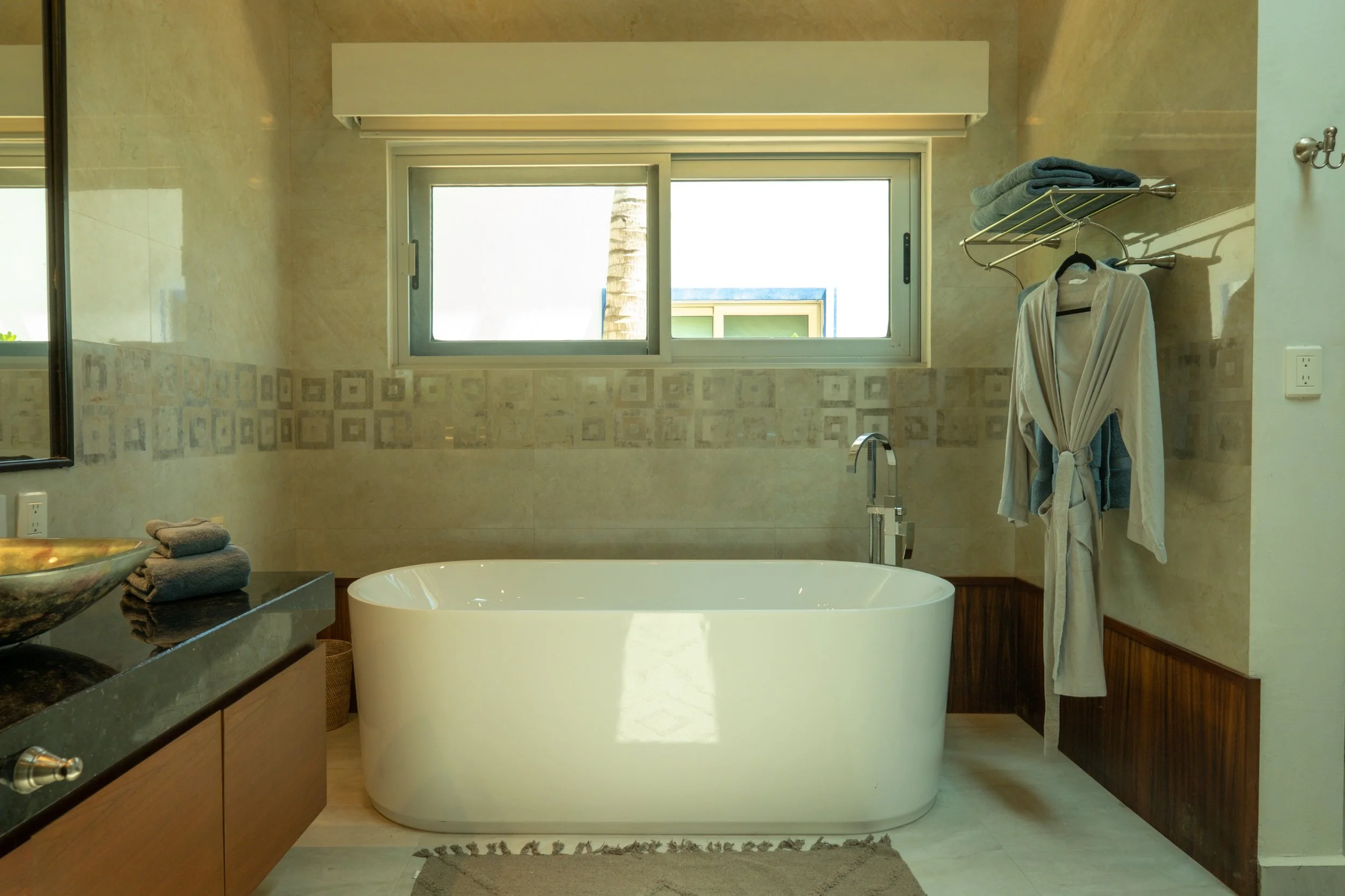 Modern bathroom with a freestanding bathtub, window above it, a countertop with towels, and a robe hanging on a wall-mounted rack with towels.