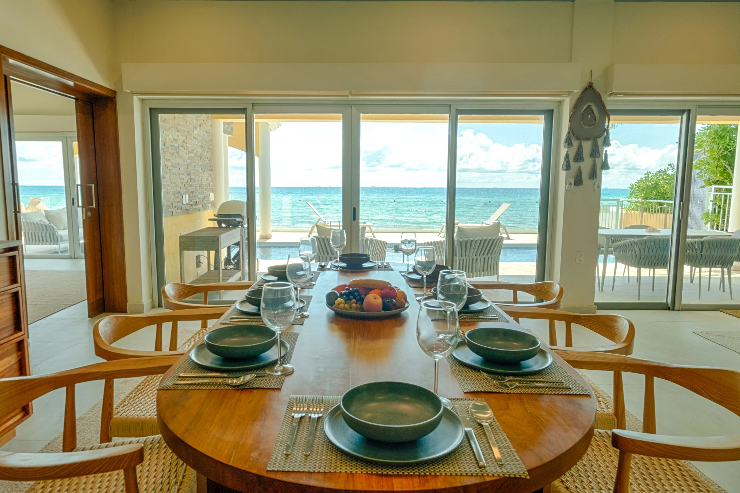 Dining room with a wooden table set with black bowls, glasses, silverware, and a fruit centerpiece, overlooking a sunny ocean view through sliding glass doors.