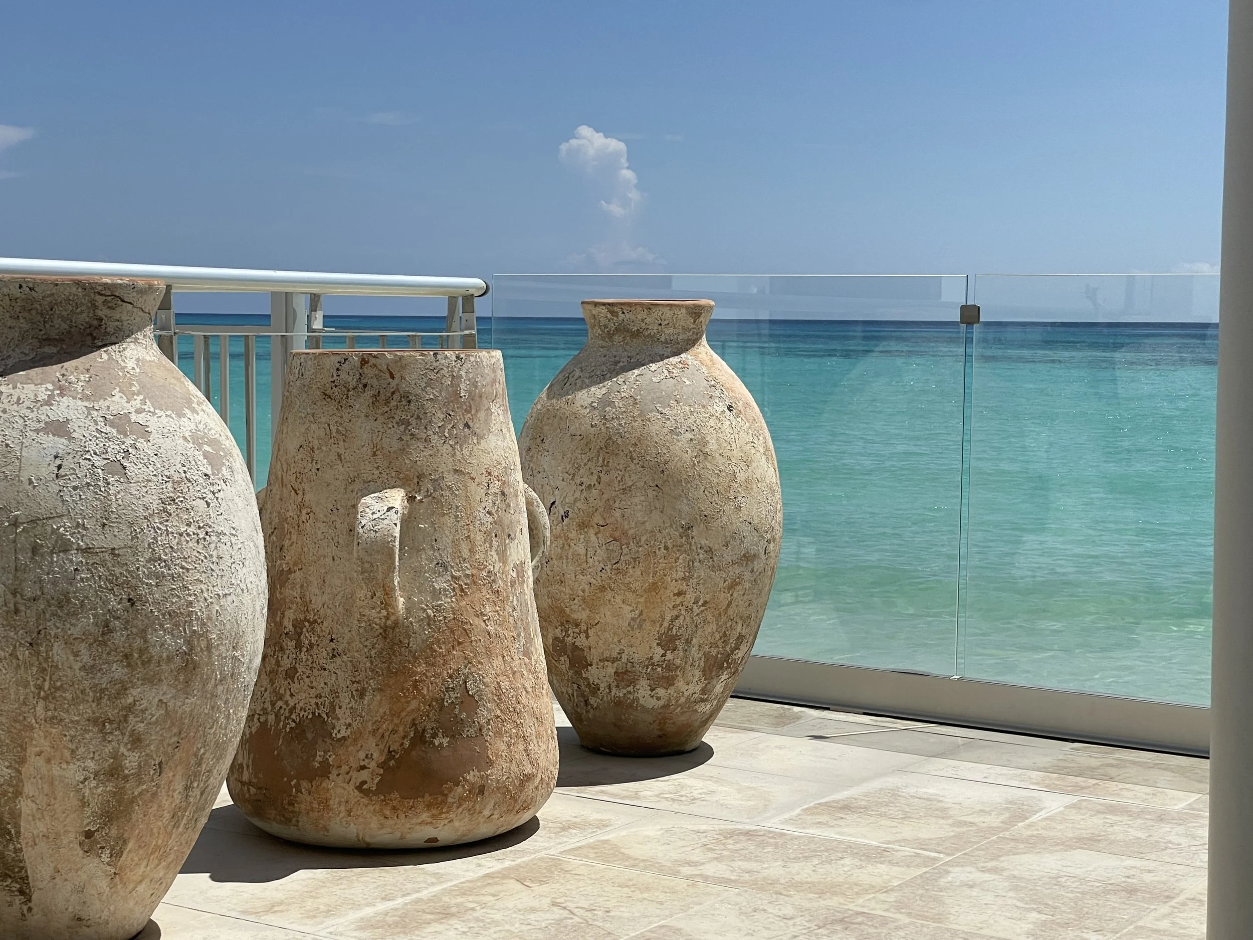 Three large, weathered clay pots on a tiled balcony overlooking the ocean, with clear turquoise water and a blue sky in the background.