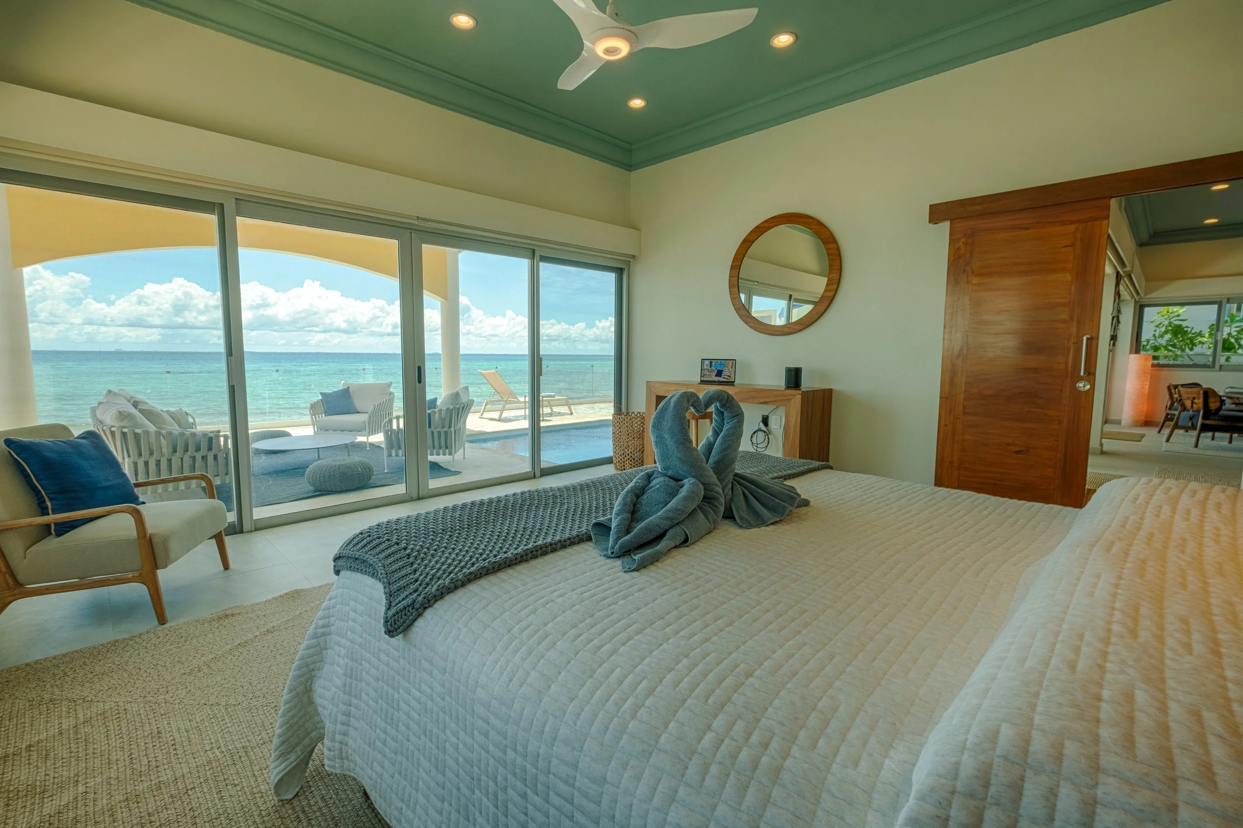 Beachfront bedroom with sliding glass doors leading to a balcony overlooking the ocean, with furniture including a bed with towel swans, an armchair with a cushion, and a view of the sea and sky.