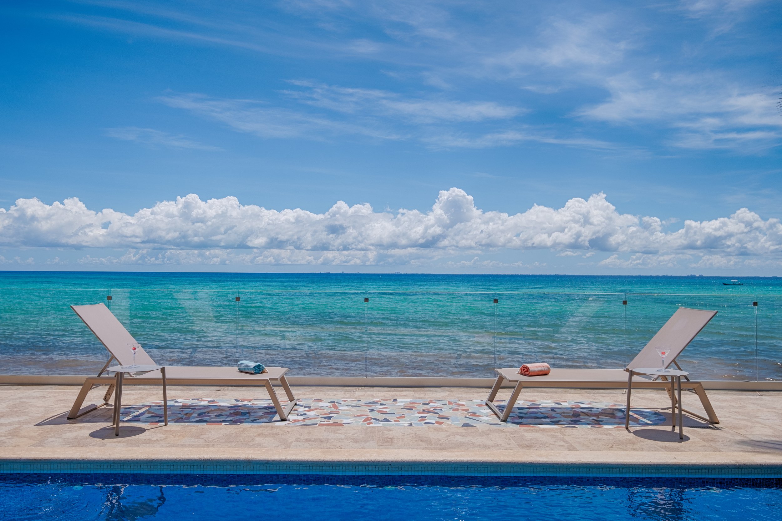 Two sun loungers and a small side table beside a swimming pool, with the ocean and cloudy sky in the background.