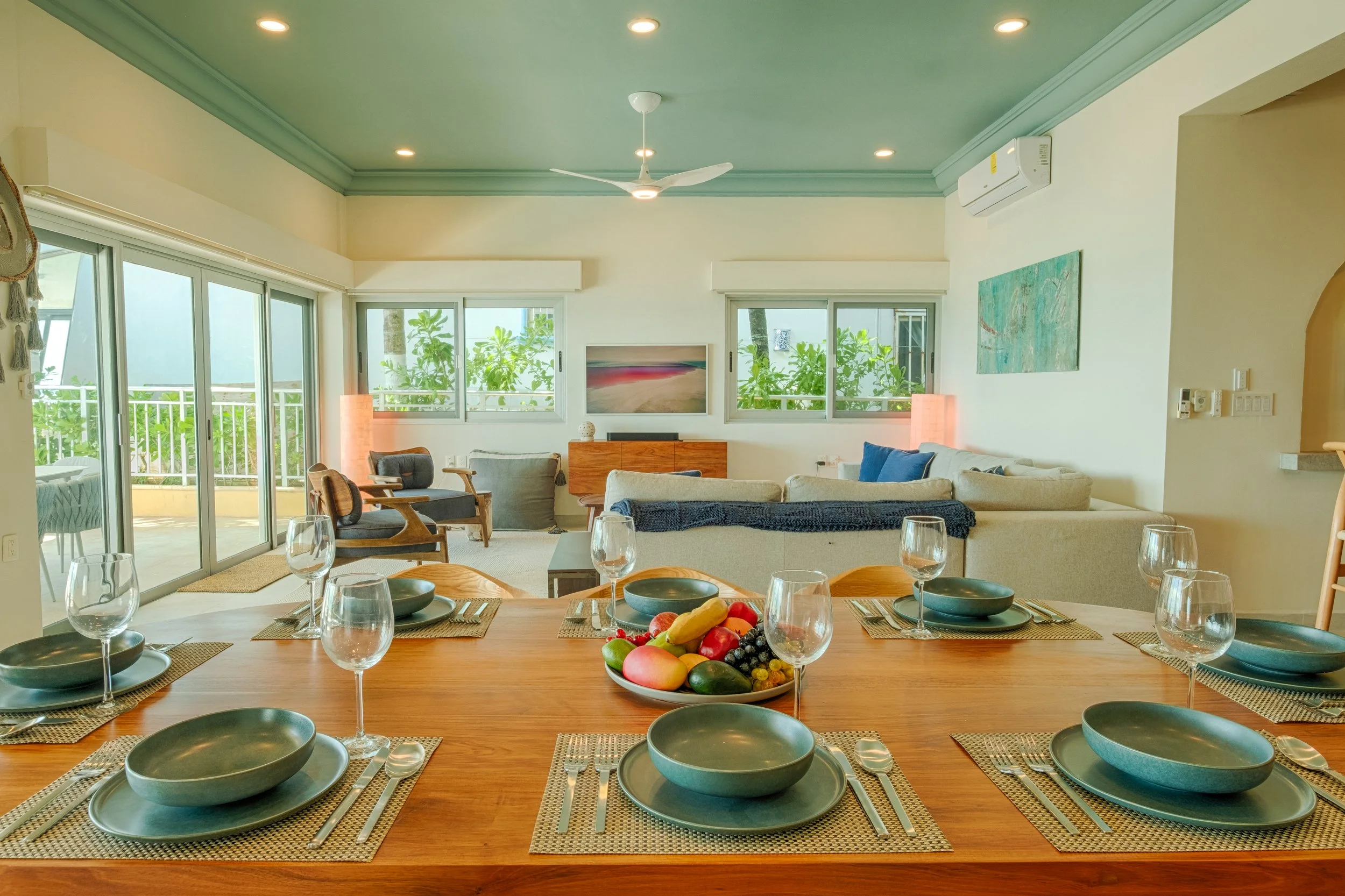 Open-concept living and dining room with a large wooden table set with six place settings, a fruit bowl, and a view into a living area with sofas, armchairs, a TV, and glass sliding doors leading to a balcony or patio.