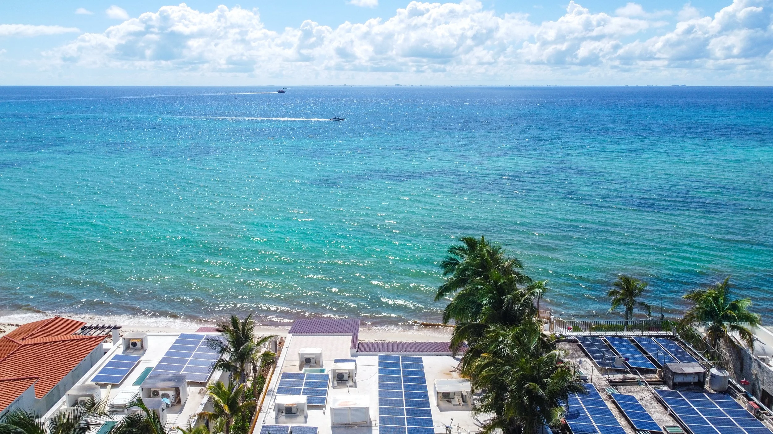 Aerial view of a tropical beach with turquoise water, palm trees, rooftops with solar panels, and boats on the horizon.