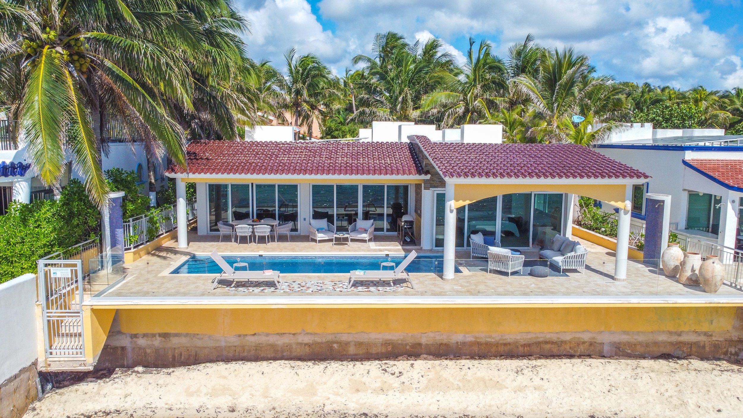 A tropical house with a swimming pool, palm trees, and outdoor furniture, including lounge chairs, chairs, and sofas, under a partly cloudy sky.