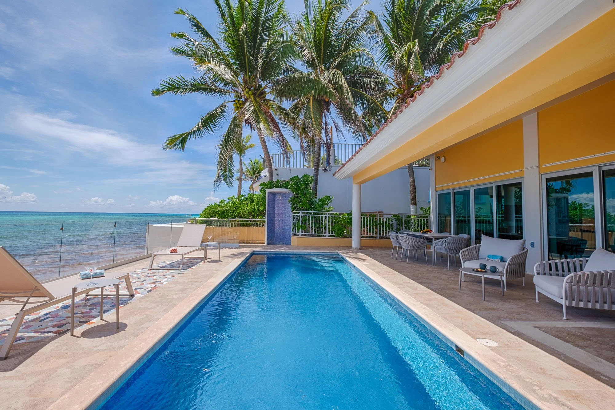 Poolside view of a backyard with a swimming pool, lounge chairs, white patio furniture, tropical palm trees, a beachfront, and an ocean in the background under a partly cloudy sky.