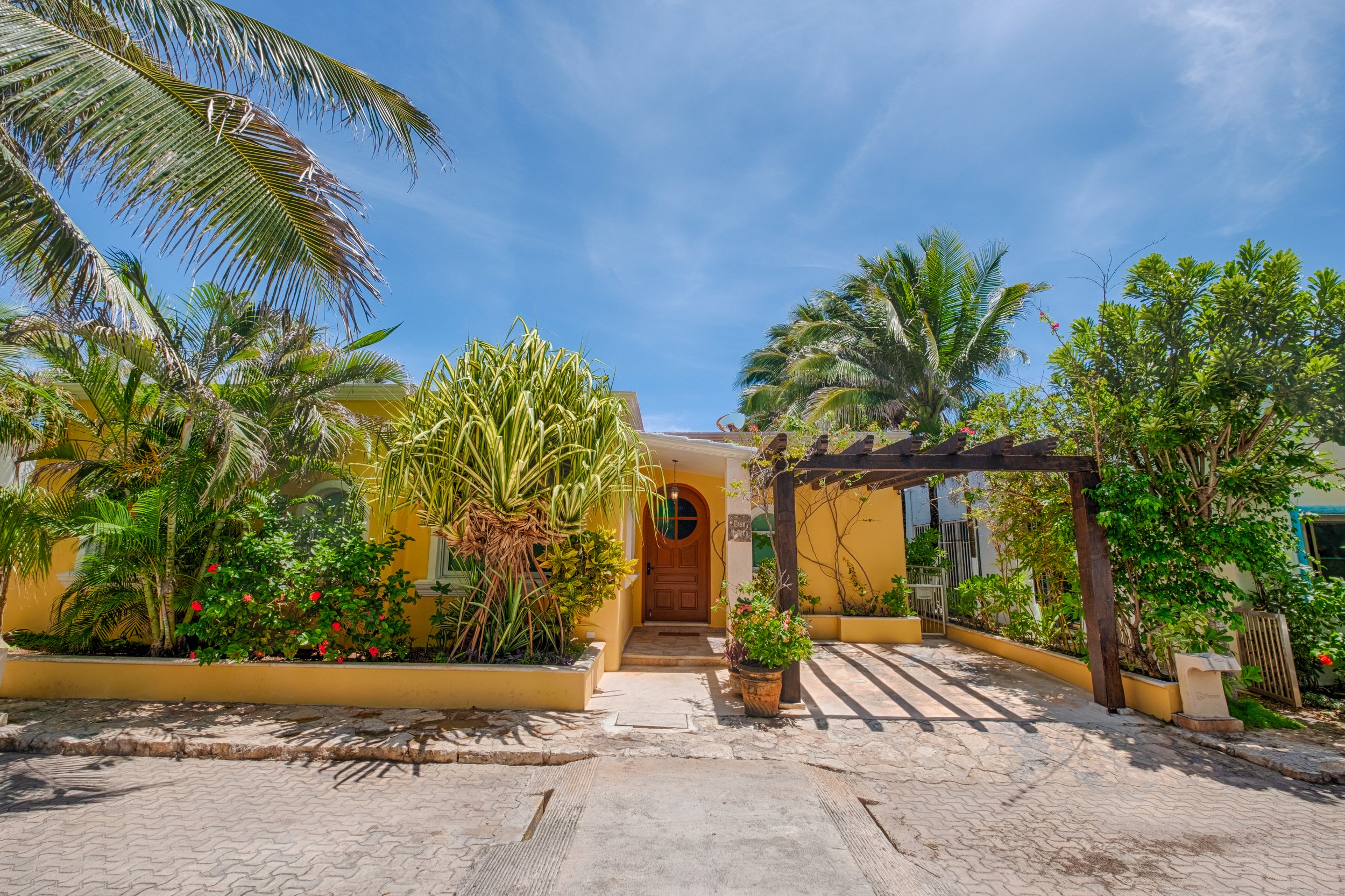 A bright yellow house with a wooden front door, surrounded by tropical plants and trees under a clear blue sky.