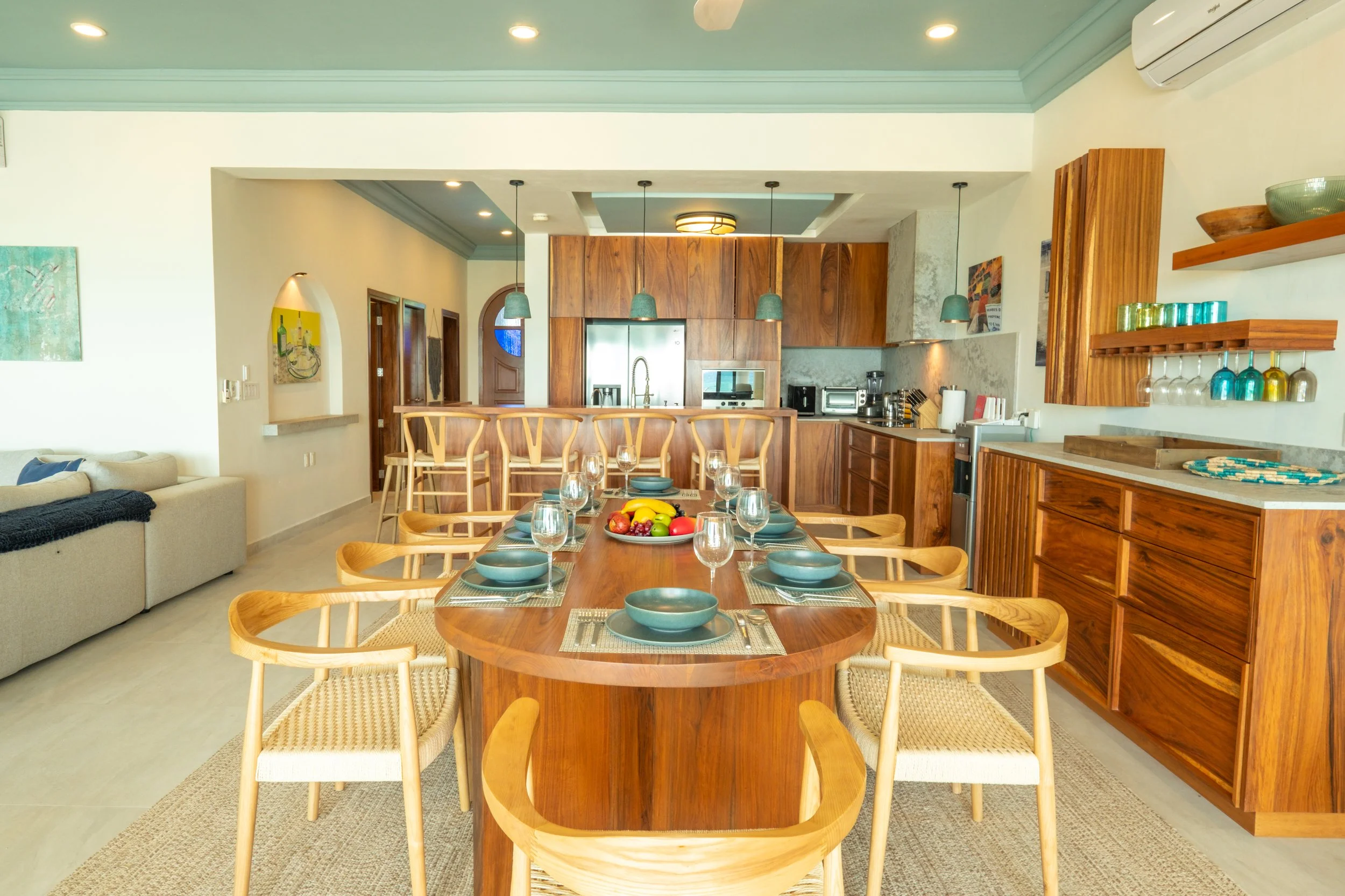 Modern open-plan kitchen and dining area with wooden furniture, a table set with blue plates and glasses, and a kitchen with wooden cabinets, stainless steel appliances, and hanging green pendant lights.