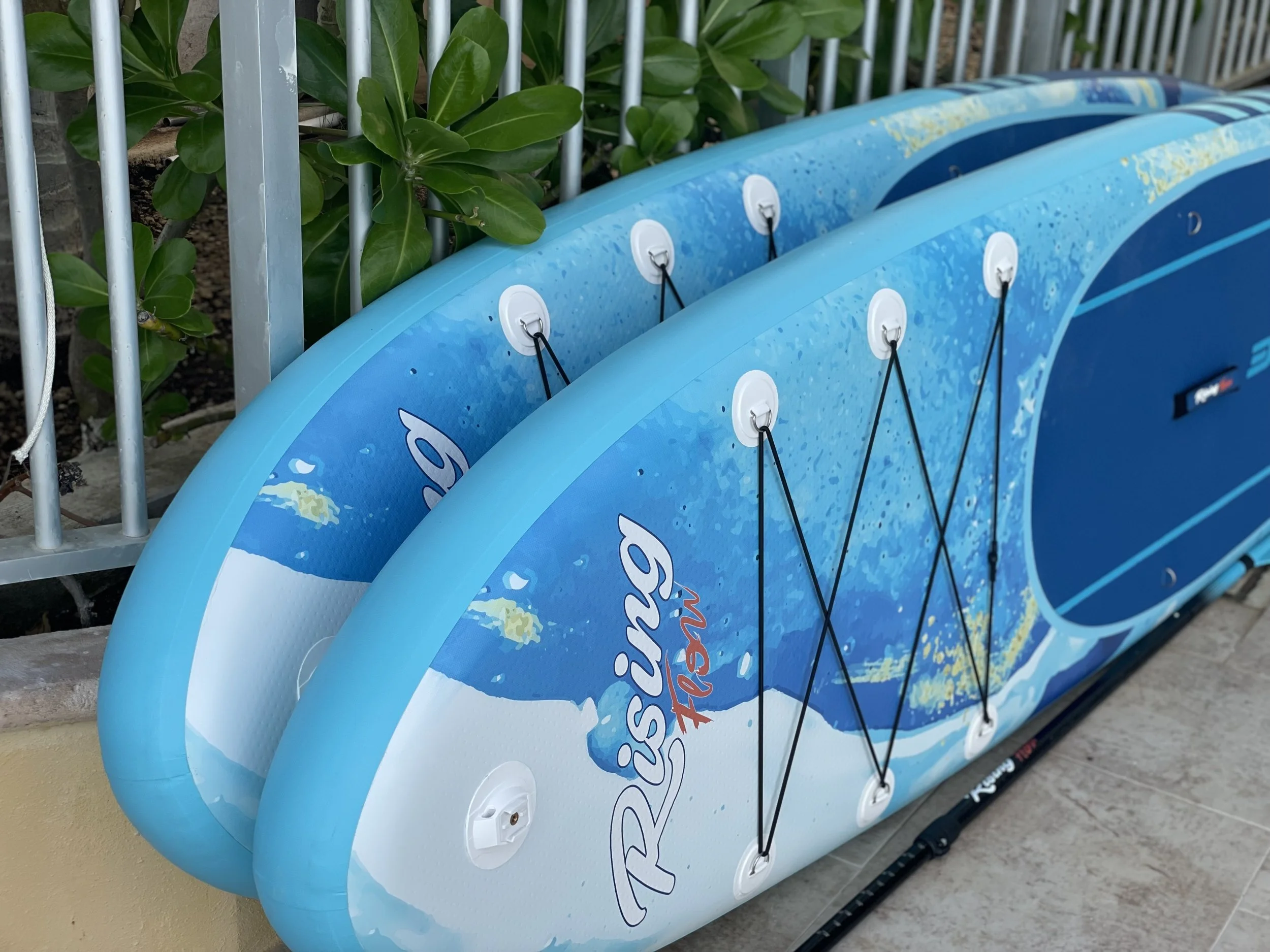 Two blue inflatable paddleboards stacked against a metal fence with green plants behind them, water droplets on the surface, and the words 'Rising Reef' printed on the side.