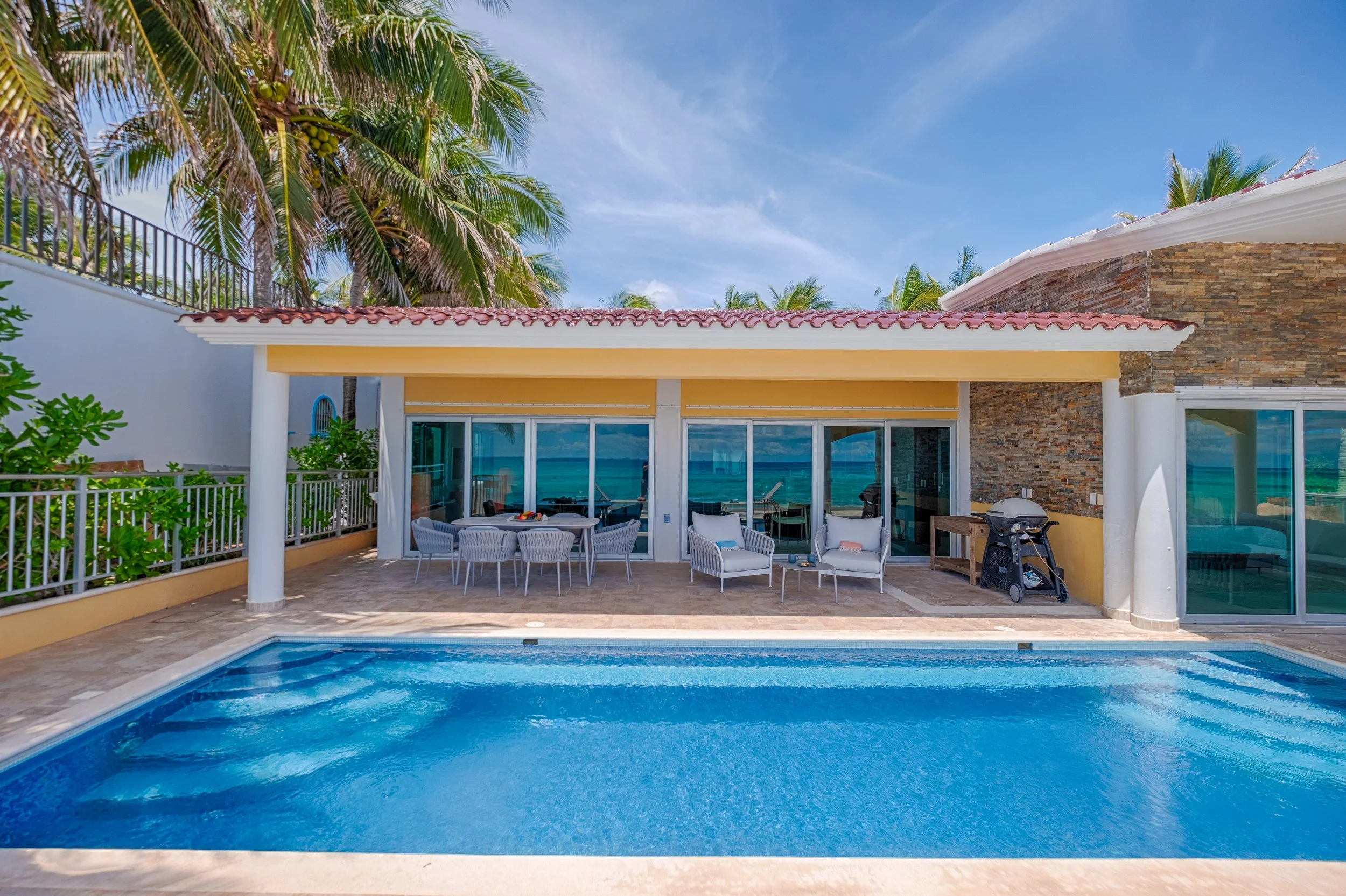 A house with a swimming pool in the backyard, surrounded by tropical palm trees, with outdoor patio furniture, a barbecue grill, and a view of the ocean in the background.
