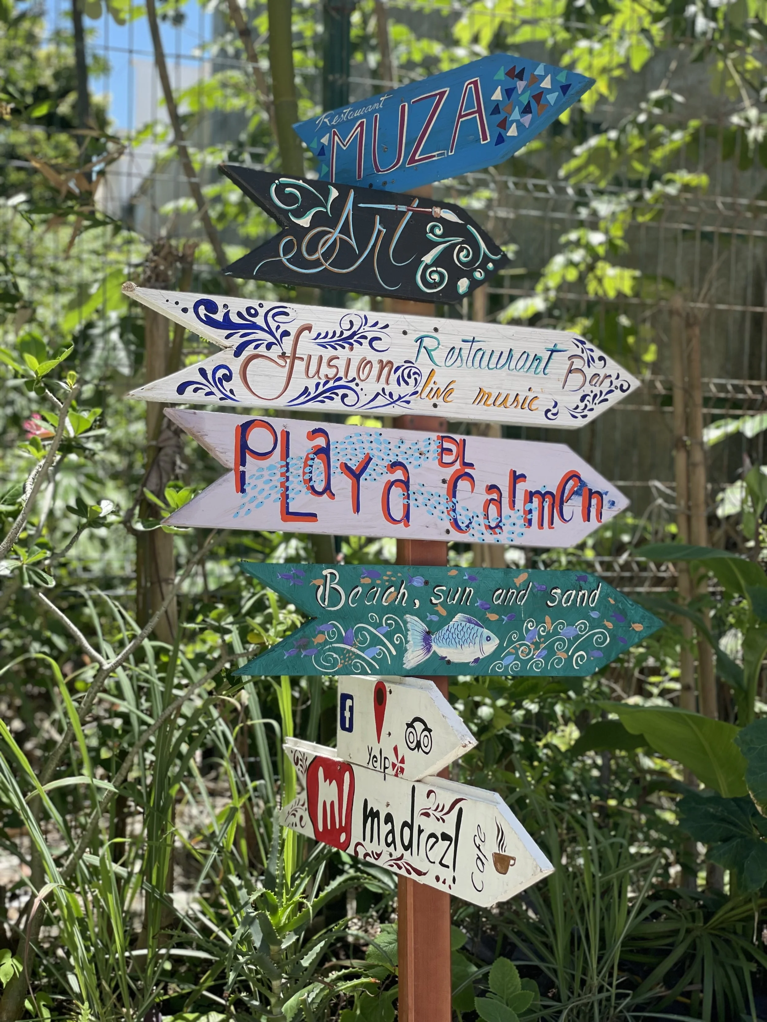 Colorful wooden directional signs pointing to various attractions and amenities, including a restaurant called Muza, an art activity, live music, Playa Carmen beach, and a place called Madreza, surrounded by green plants and foliage.