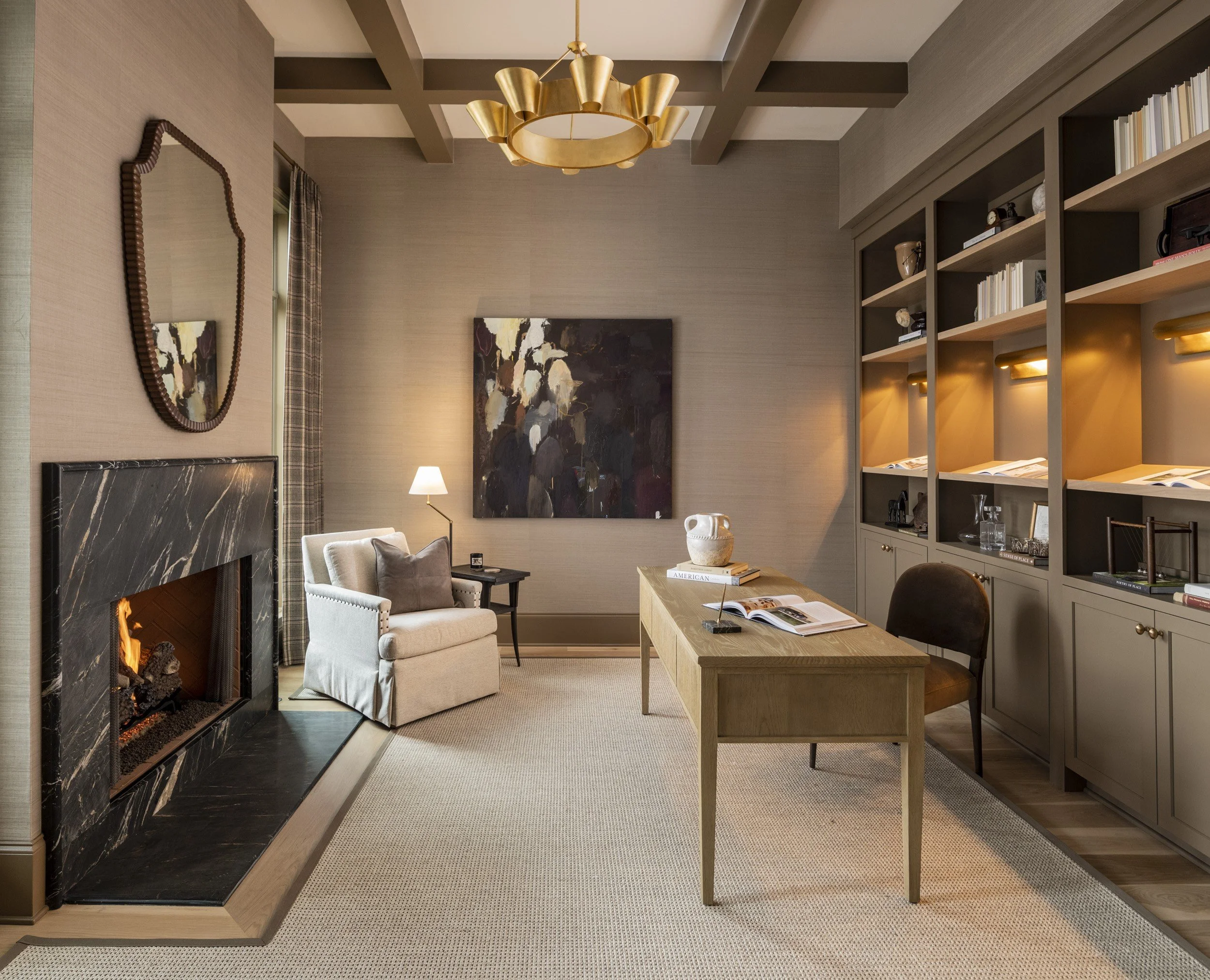 Living room with gray walls, fireplace with black marble, beige armchair, wooden coffee table, built-in bookshelves with books and decor, abstract painting, table lamp, and gold chandelier.