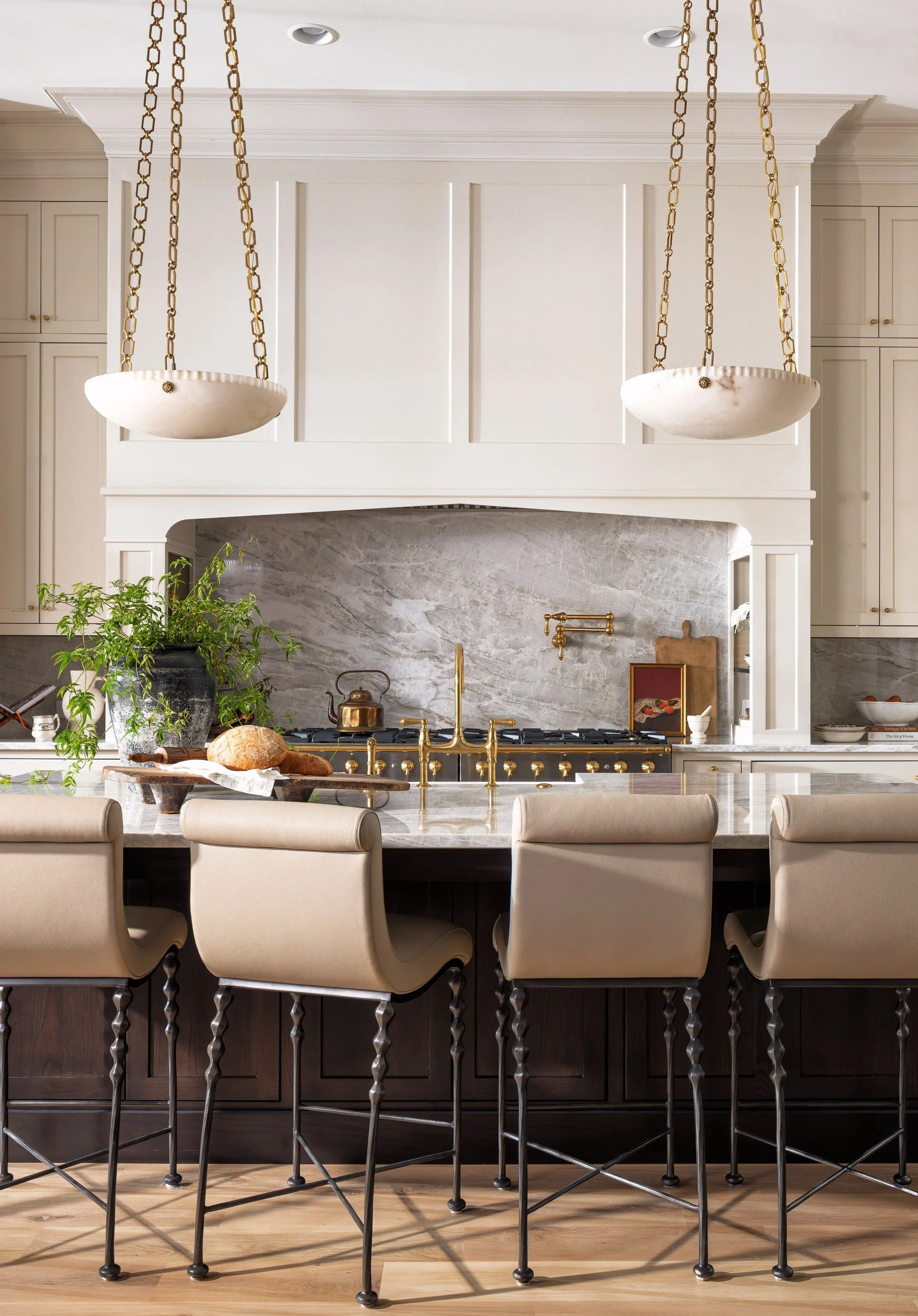 A kitchen with a marble island, beige chairs, a large potted plant, and a backsplash with a marble pattern. Two large hanging light fixtures with chains are above the island.