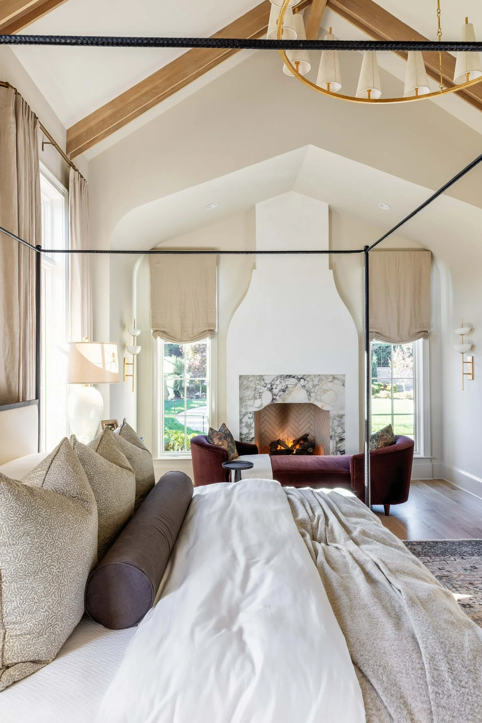 A cozy living room with a white ceiling and large windows with beige curtains, a gas fireplace with a marble surround, a purple velvet sofa, and a bed with beige patterned pillows in the foreground. A gold chandelier hangs from the ceiling.