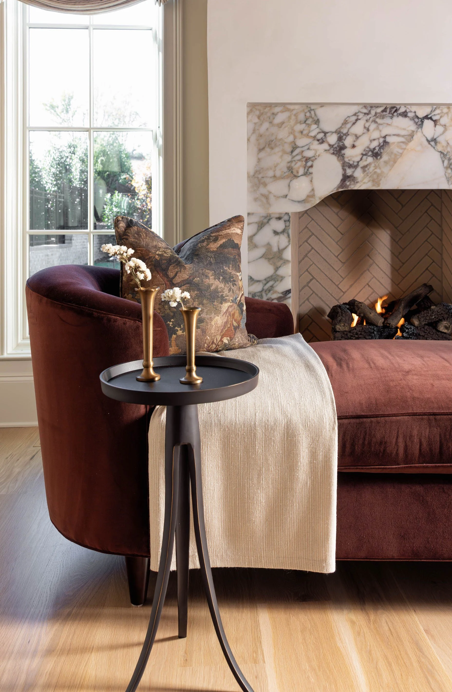 A cozy living room corner featuring a burgundy velvet armchair with a beige cushion, a small black side table with two gold candle holders, a window with white trim letting in natural light, a white wall with a small fireplace with a marble mantel and a brick interior, and a fire burning inside the fireplace.