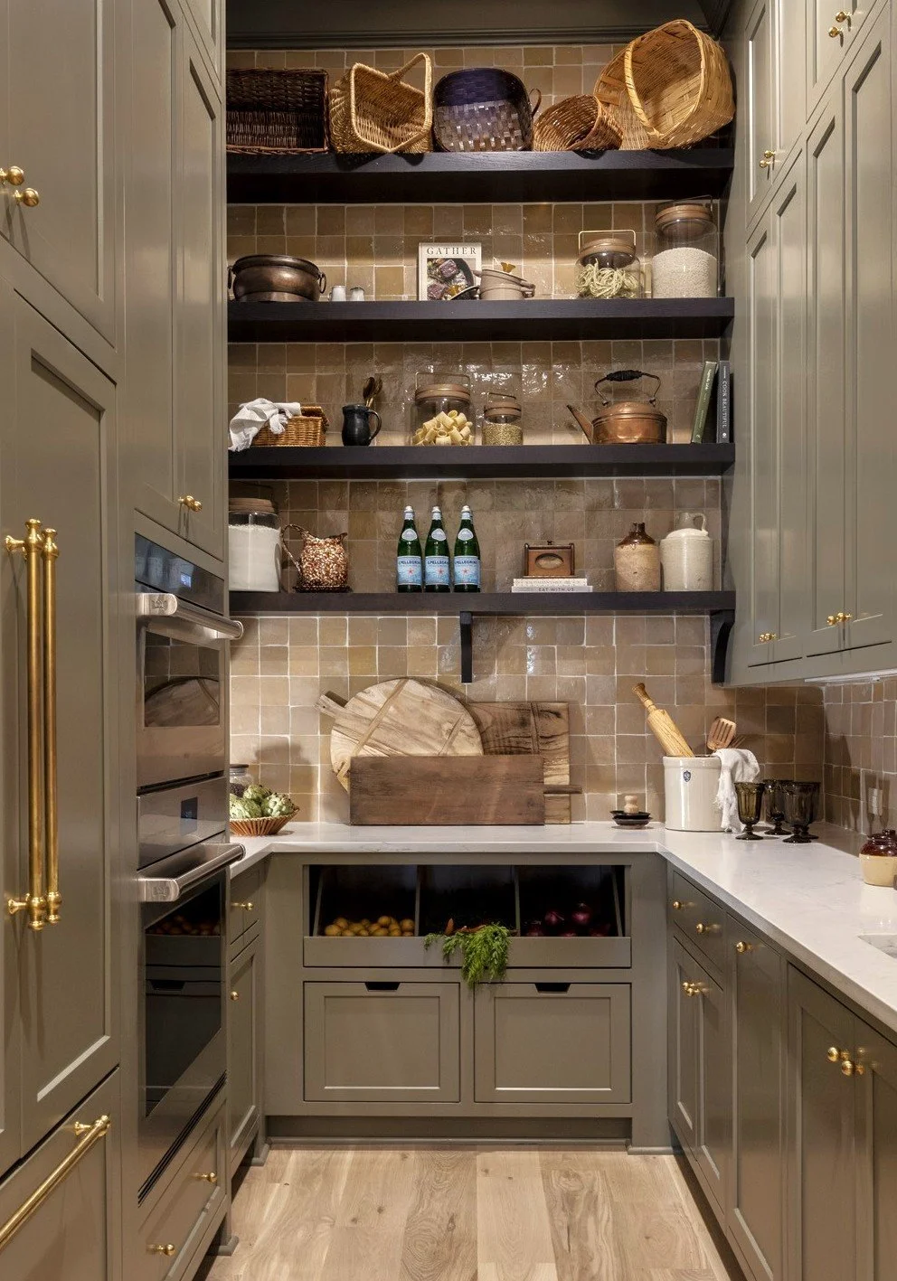A cozy kitchen with green cabinets, open shelving, and a beige tiled backsplash. The shelves hold woven baskets, jars, and kitchen items. The countertop has cutting boards, a white container with utensils, and some dark glasses. Below, open compartme
