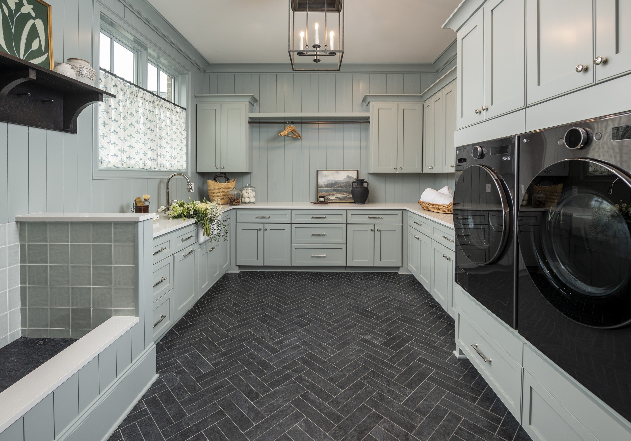 A laundry room featuring light gray cabinetry, a black and silver front-load washer and dryer, a window with patterned curtains, a black wall-mounted shelf with vases, a hanging light fixture, a countertop with decorative jars, a framed painting, a b