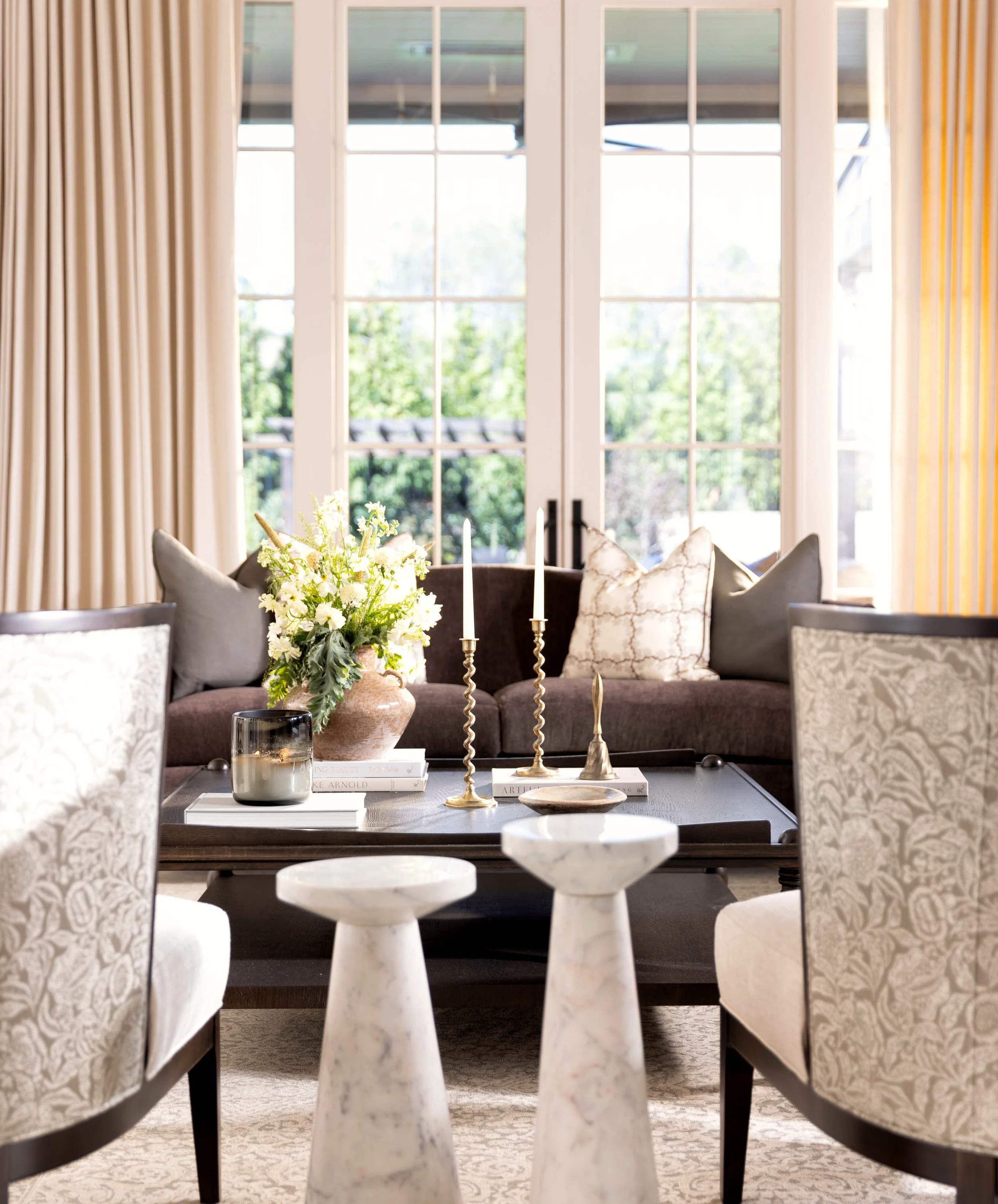 A living room with large glass doors, beige curtains, a dark sofa with patterned pillows, a coffee table with decorative objects, and a chandelier. There is greenery outside.