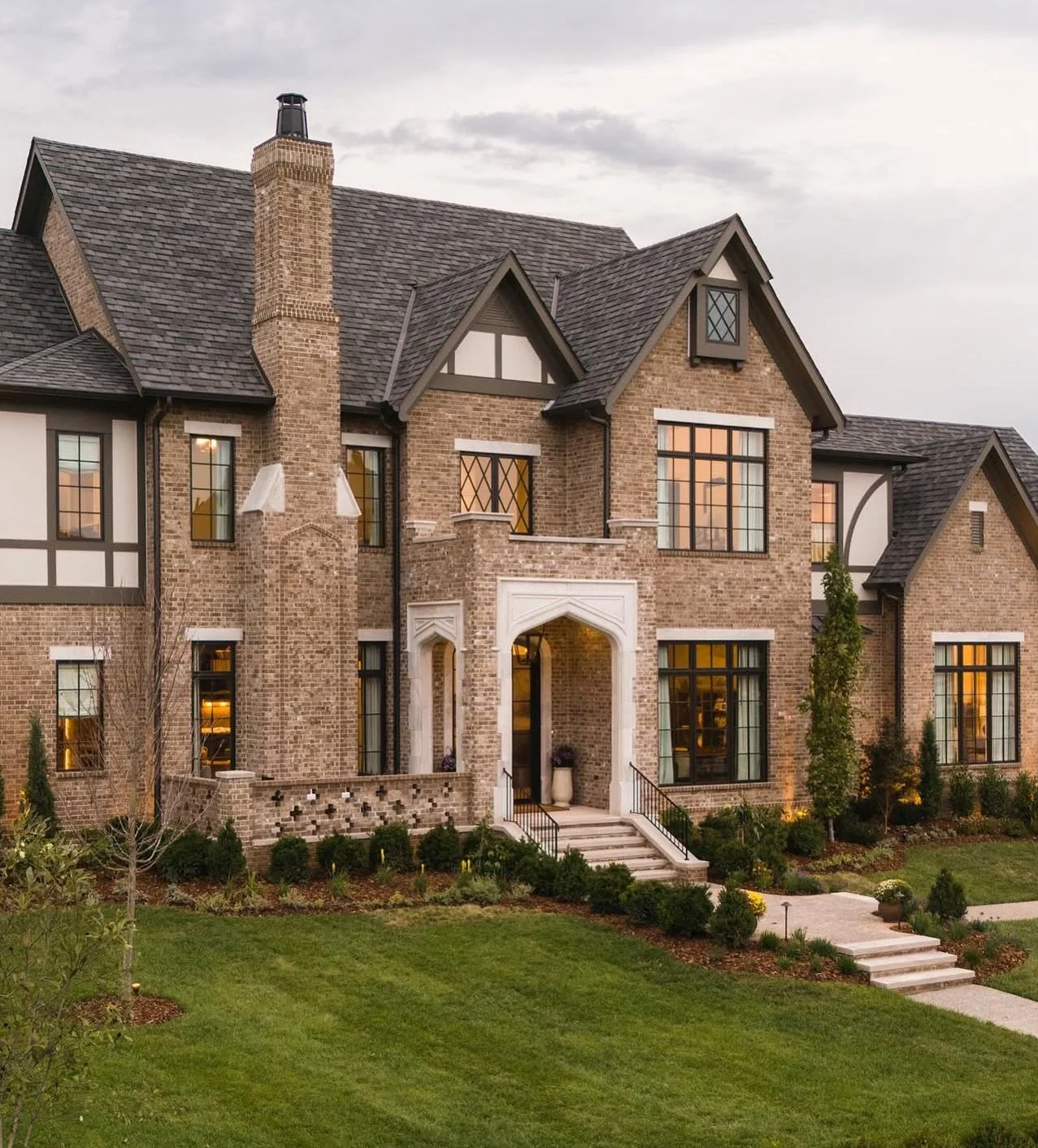 Large brick house with multiple gabled roofs, tall chimney, and decorative windows, surrounded by a landscaped lawn with bushes and small trees.