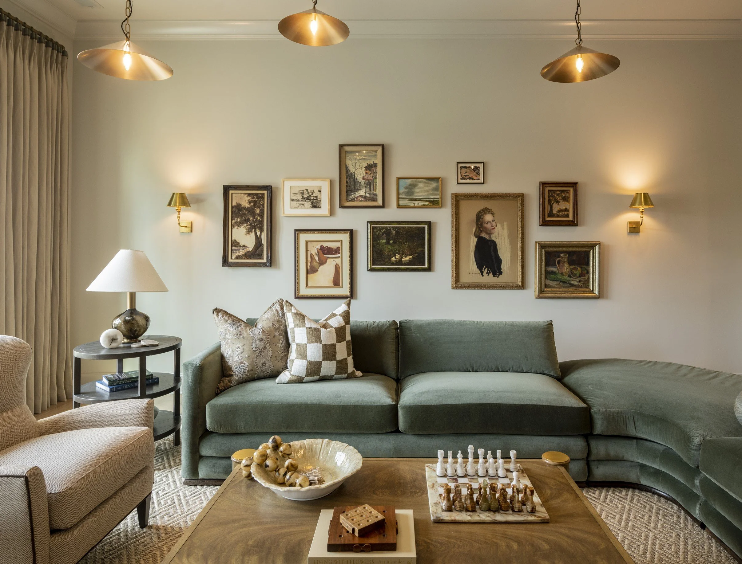 Living room with a green sofa, patterned pillows, a wooden coffee table with a chess set, a decorative bowl with balls, a beige armchair, a side table with books and a lamp, framed artwork on the wall, and gold pendant lights hanging from the ceiling