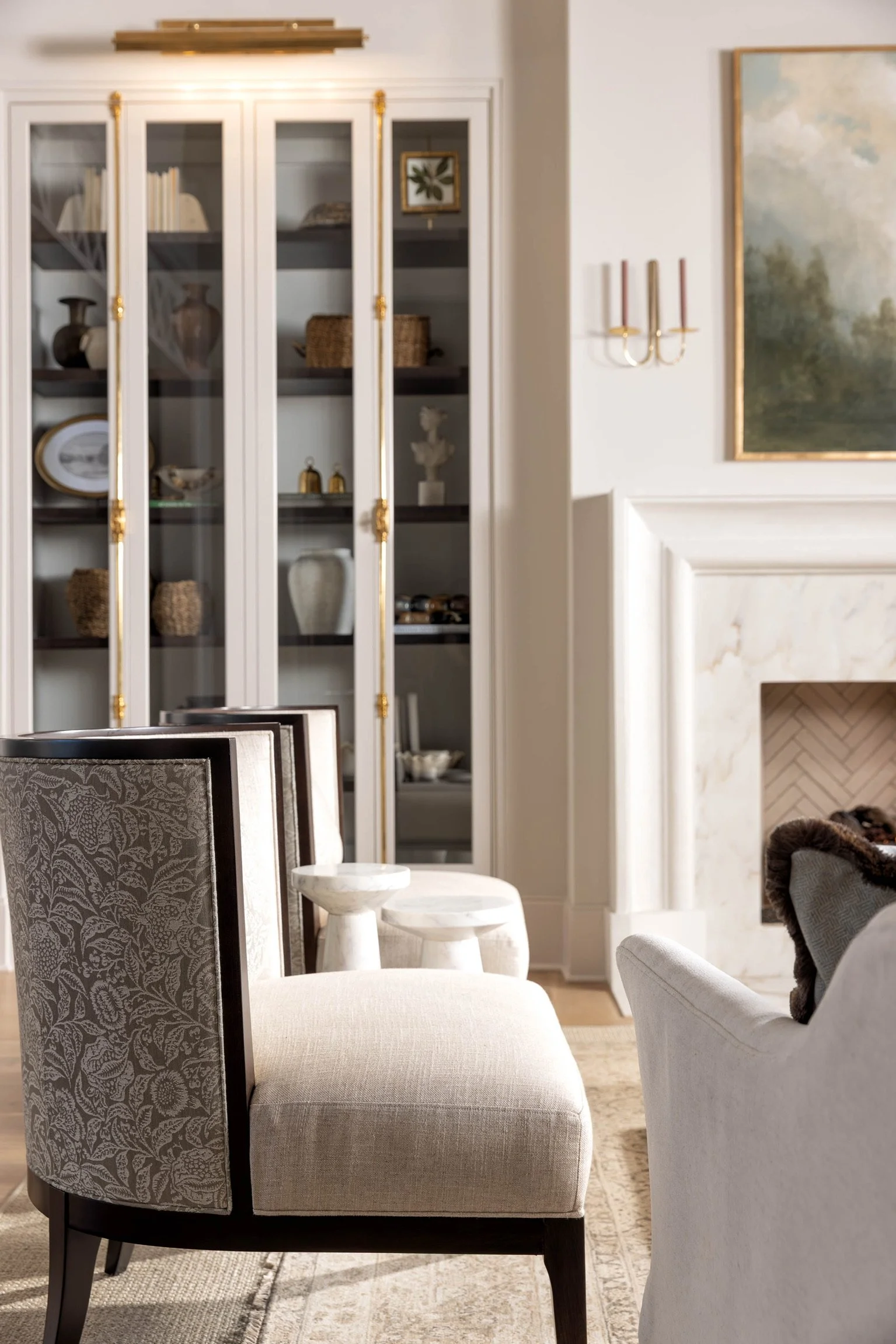 A living room with a built-in white cabinet displaying decorative items, a wall-mounted candle holder, a framed painting, a white marble fireplace, and upholstered chairs with patterned fabric.