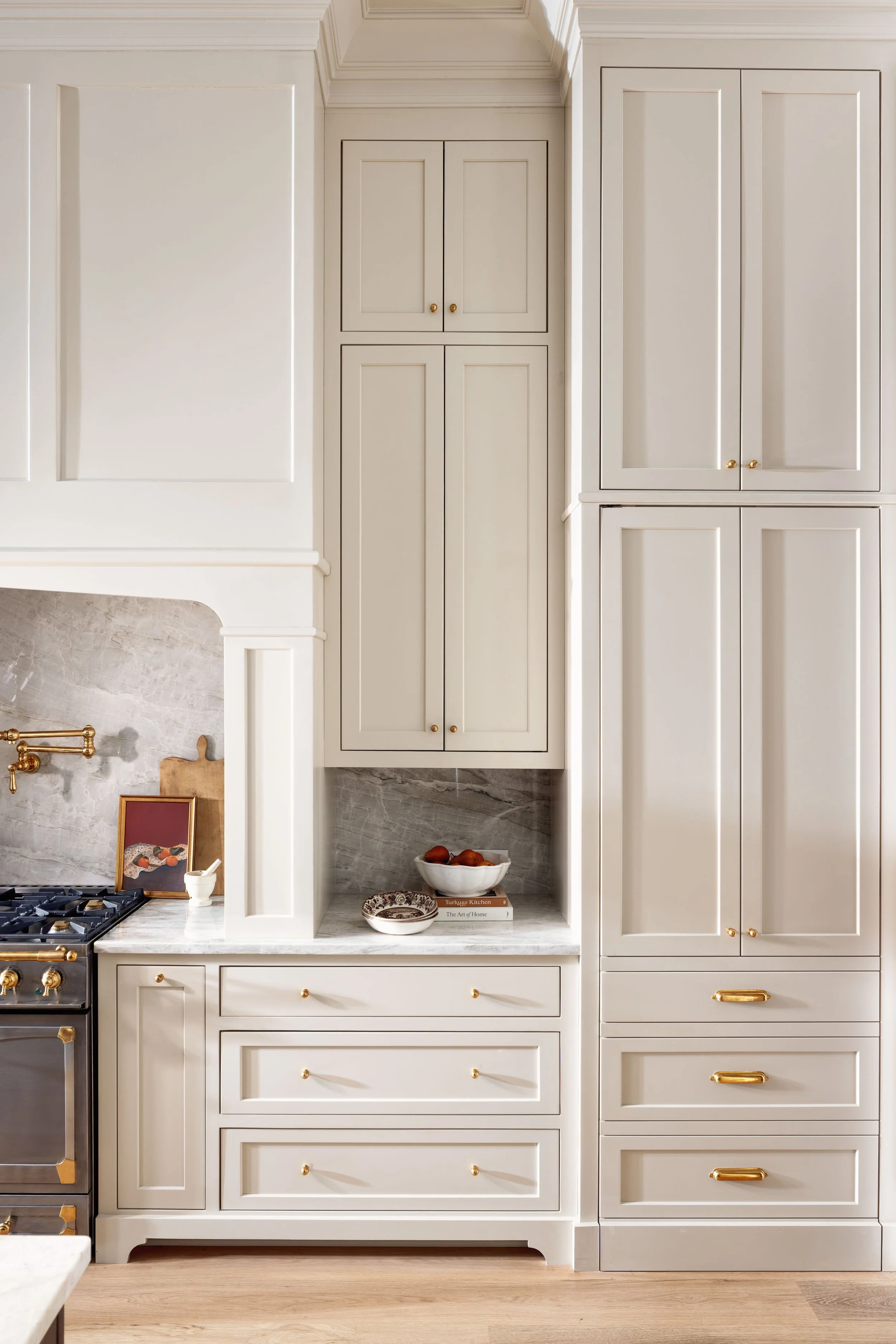 A kitchen with white cabinetry, gold handles, and a marble countertop and backsplash. Open shelves hold bowls and cookbooks, with a decorative fireplace and picture frame on the side.