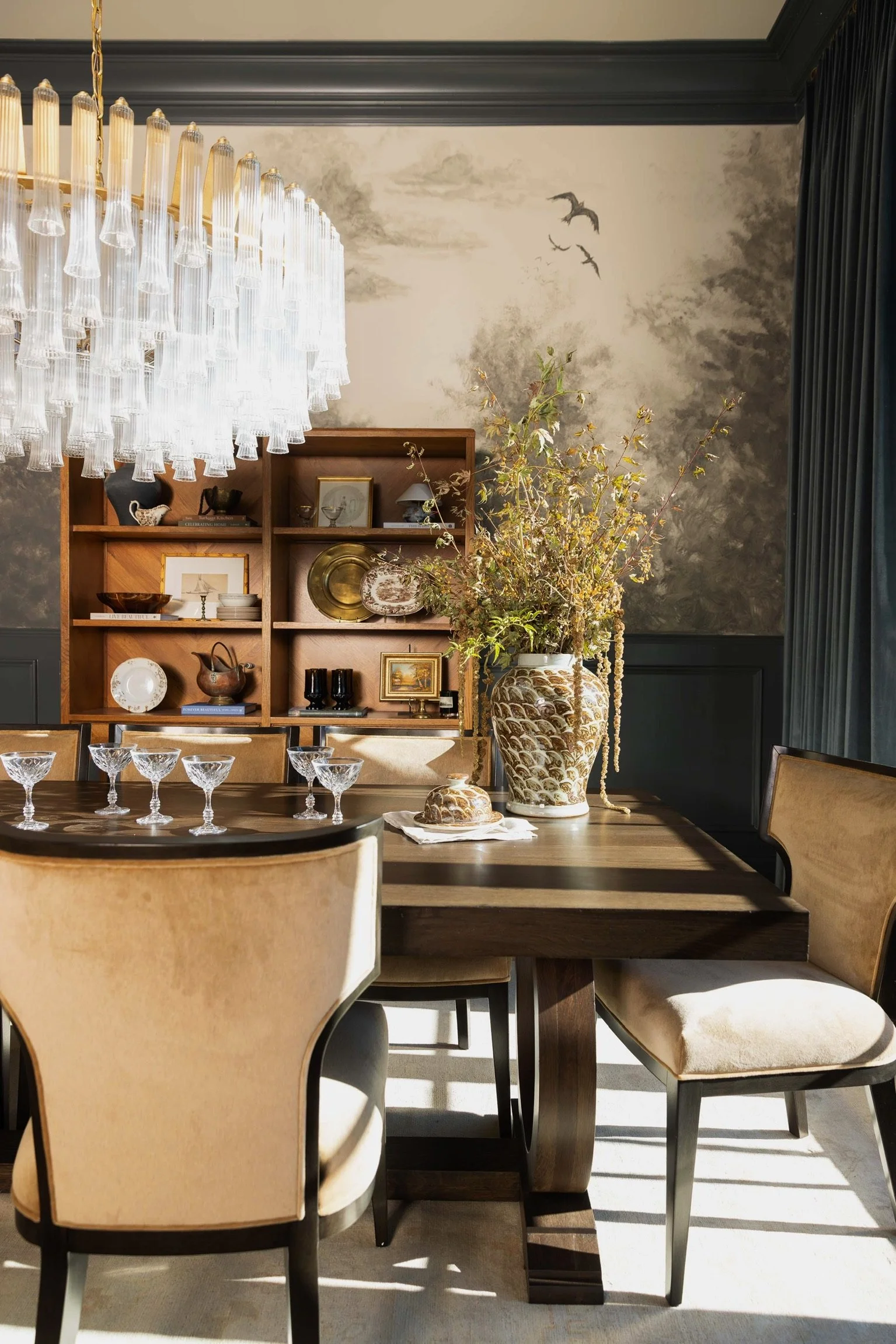 A dining room with a wooden table, beige upholstered chairs, a large floral arrangement in a decorative vase, a chandelier, and a sideboard with various decorative items and framed pictures. The wall features a cloud and bird mural.