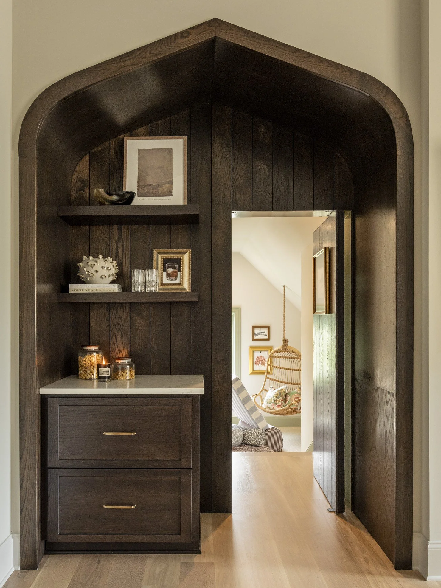 Dark wooden archway with built-in side shelves, decorated with framed artwork, a black ceramic bowl, and books. Visible room beyond with hanging rattan chair, patterned pillows, and framed wall art, hardwood flooring.