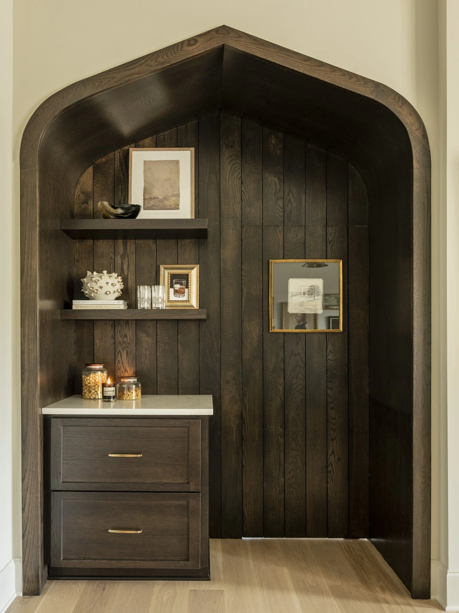 A built-in corner shelf and cabinet with dark wood finish, decorated with framed artwork, glassware, a ceramic piece, jars of snacks, and candles on a white surface, with a light-colored wooden floor.