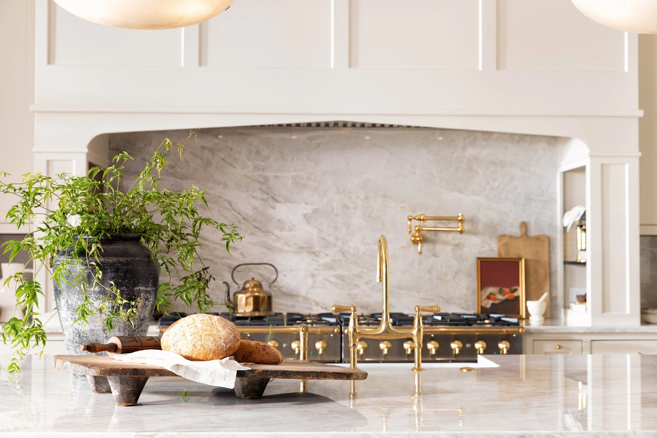 A marble kitchen island with a wooden cutting board, fresh bread, and a black vase with green plants. The background features a white and gray marble backsplash, gold kitchen faucet, and white cabinetry.