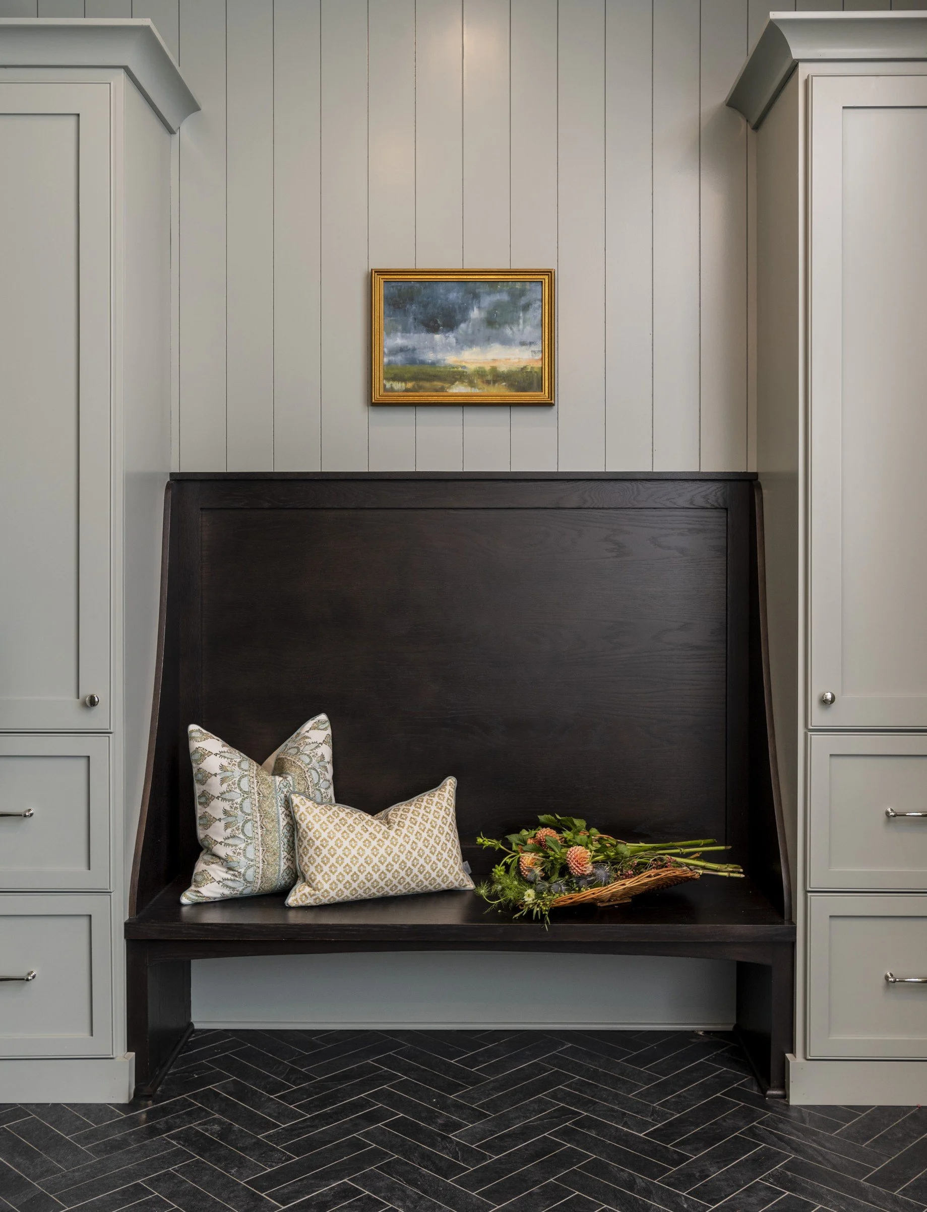 A dark wooden headboard with built-in seating area, decorated with patterned pillows, and a bouquet of flowers on the right side. The wall behind is painted in a light gray color with vertical panels, and there is a small landscape painting in a gold