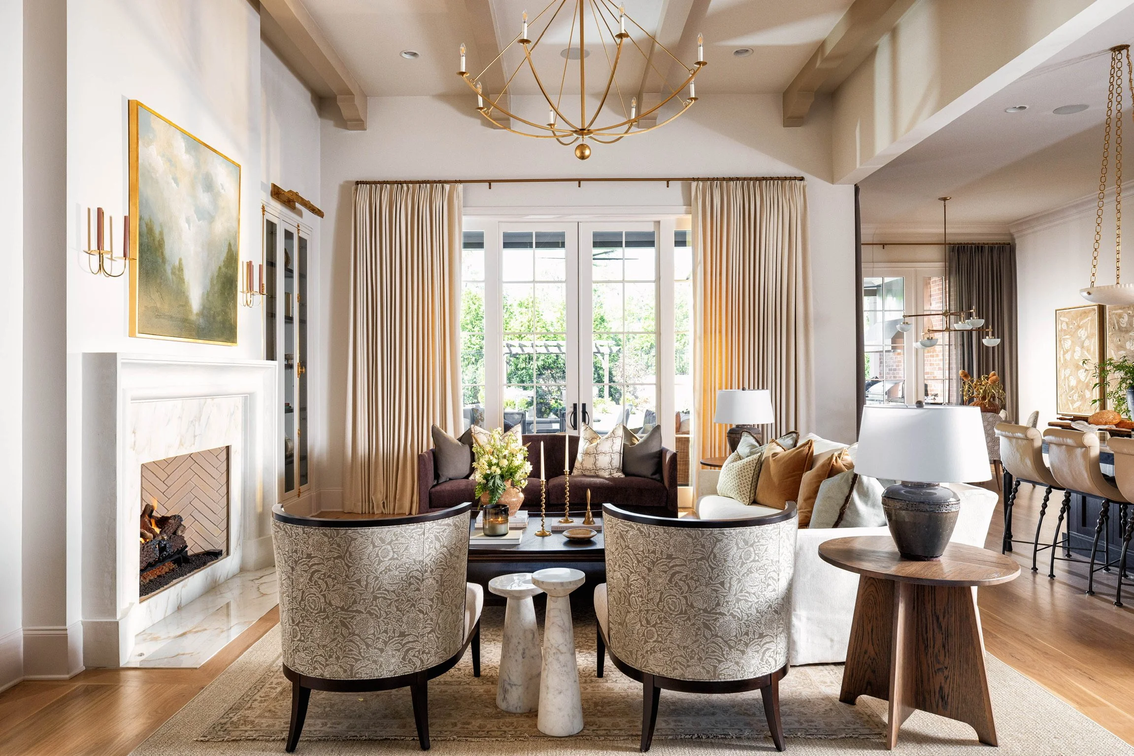 A spacious, bright living room with large windows, cream curtains, a fireplace, and elegant furniture including a round coffee table, upholstered chairs, and a variety of decorative items.