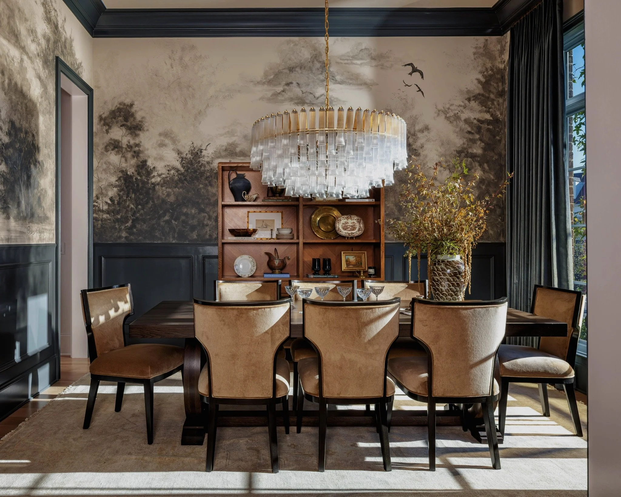 A dining room with a wooden dining table and six upholstered chairs, a large chandelier with glass pieces hanging, a wooden bookshelf with decorative items and books, and a tall plant in a decorative vase. The wall has a scenic mural with trees and b