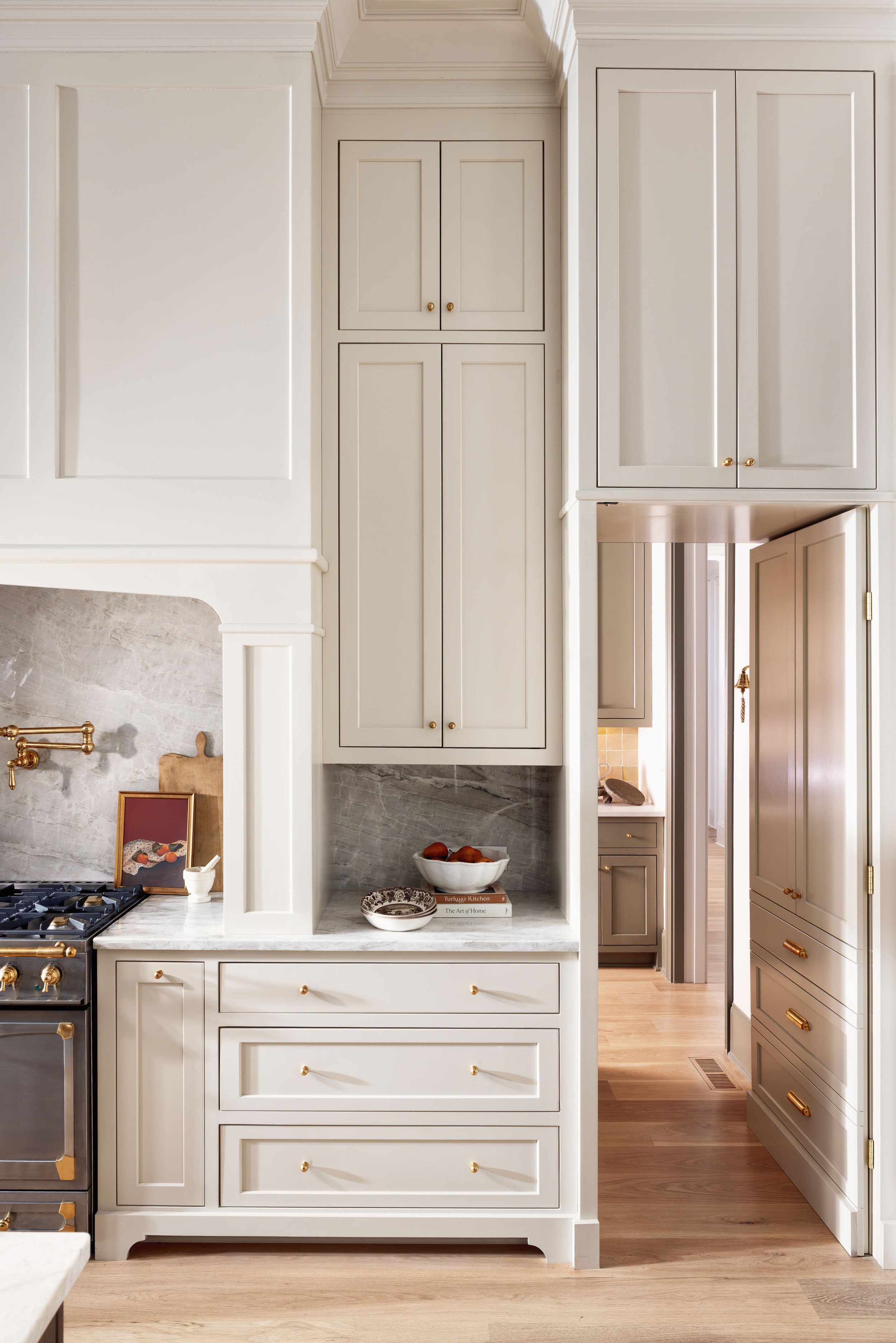 Close-up of a white kitchen showing cabinets, drawers, and part of a stove with gold accents, a marble countertop, a bowl of fruit, and a decorative plate on the counter.