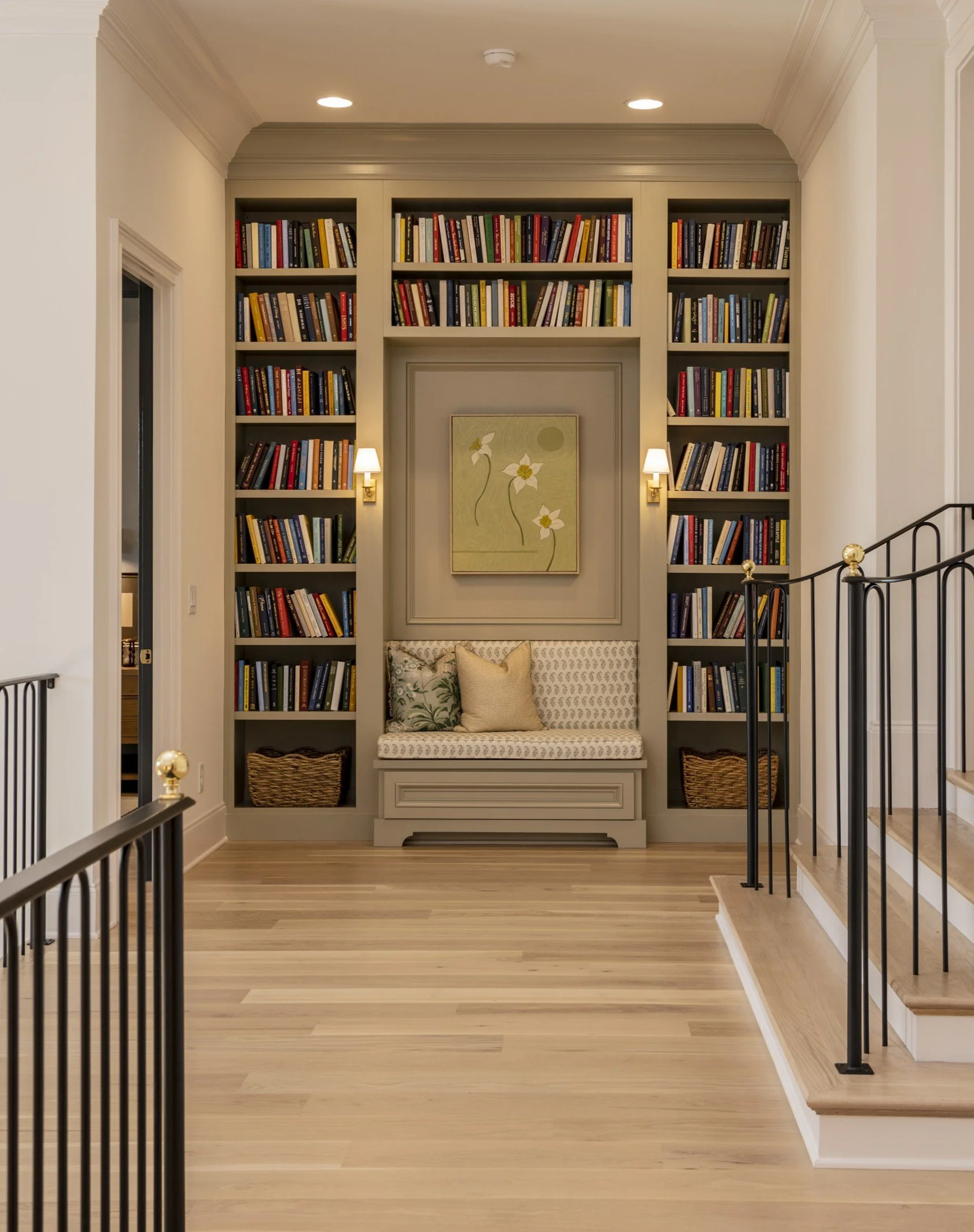 Interior of a home library with built-in bookshelves filled with books, a bench with decorative pillows, a framed floral painting, and staircase with black railings.